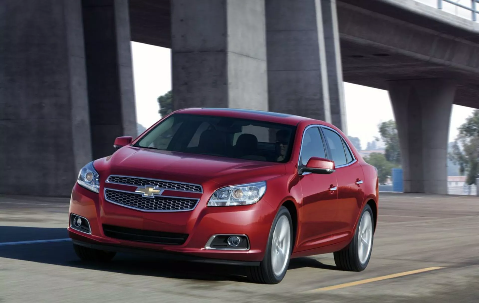 Dynamiczny widok czerwonego sedana Chevrolet Malibu 2012 w ruchu na moście, prezentujący przednią część z chromowaną kratką, smukłymi reflektorami, sportową sylwetką i aluminiowymi kołami.