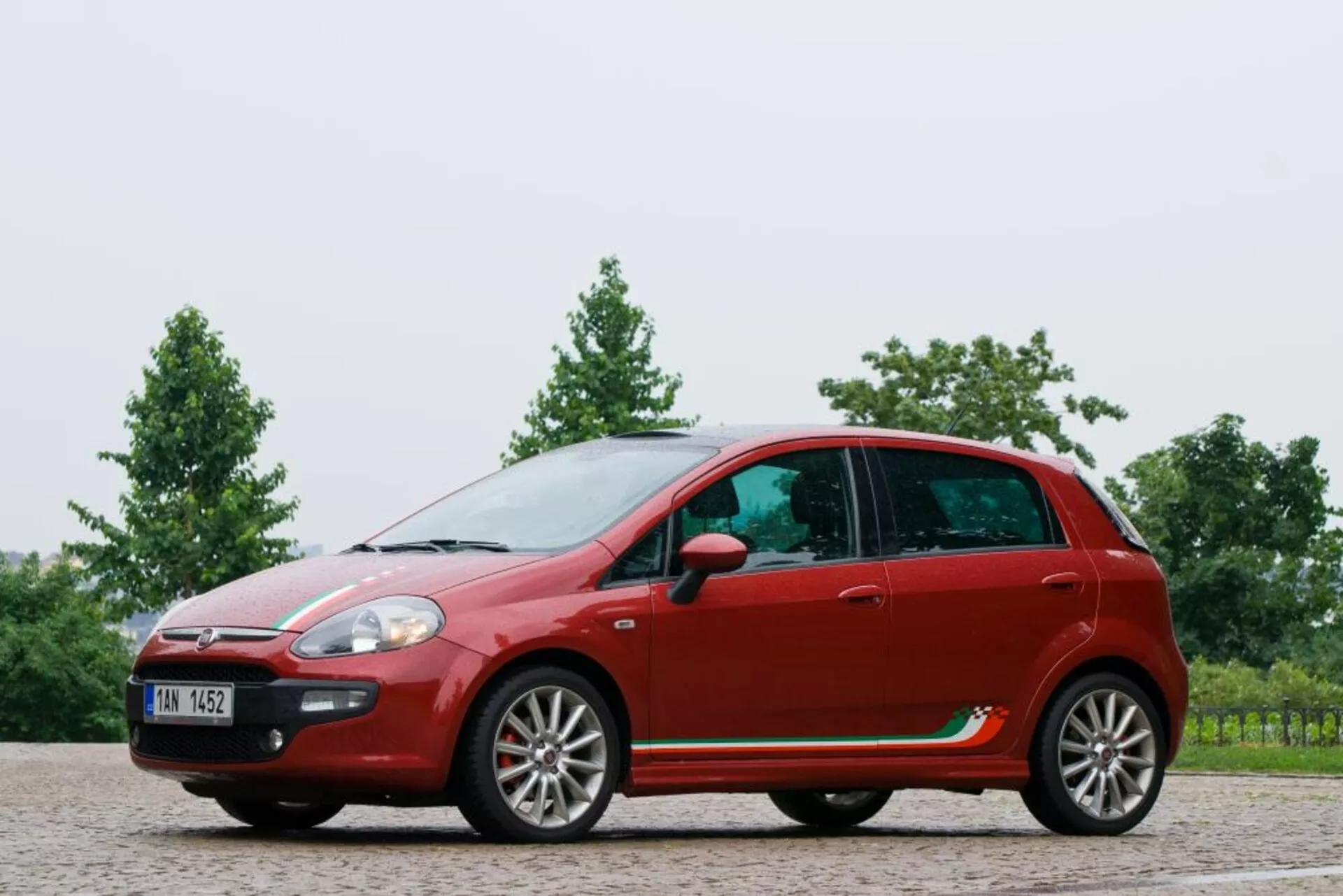 Pełny widok bocznego profilu czerwonego hatchbacka Fiat Punto z 2011 roku, pokazujący zakrzywioną linię dachu, konfigurację pięciodrzwiową z felgami aluminiowymi, lusterka boczne na drzwiach, wyraźne linie charakterystyczne i plastikowe osłony na żwirowej powierzchni z tłem zieleni.