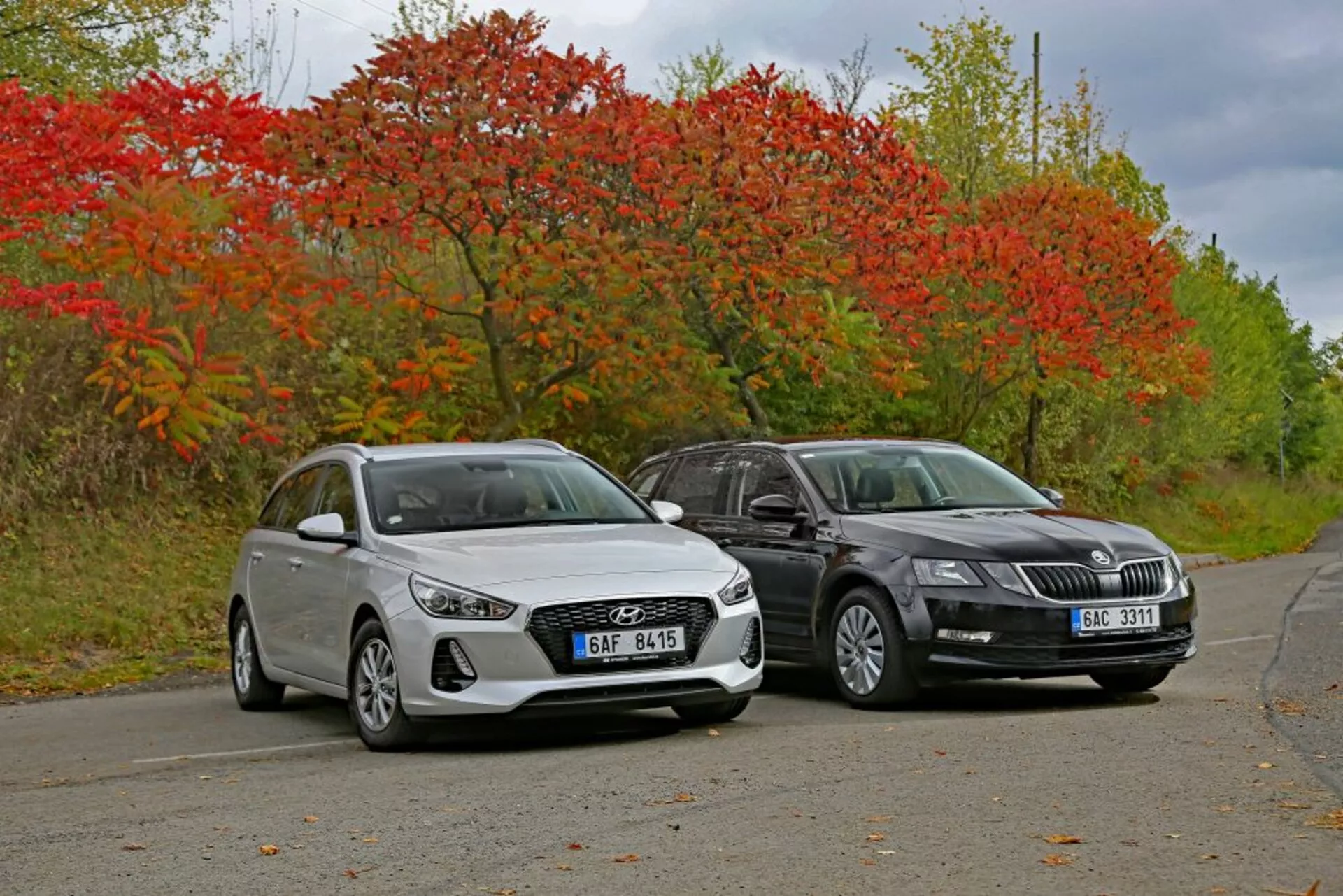 Biały hatchback Hyundai i30 z 2017 roku i ciemniejszy sedan obok siebie na cichej drodze otoczonej jesienną roślinnością, prezentujące linie wzornicze i nowoczesną technologię oświetleniową