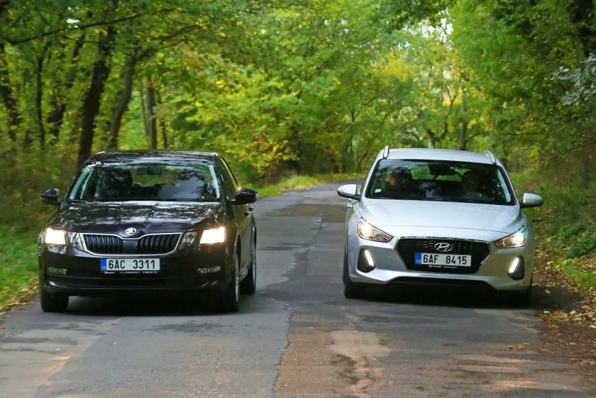 Dwa samochody stojące naprzeciwko siebie na drzewami obsadzonej wiejskiej drodze na początku jesieni: ciemnokolorowa Skoda z prostokątnym grillem i standardowymi reflektorami po lewej stronie oraz jasny Hyundai i30-N z zaokrąglonym przodem, nowoczesnymi reflektorami projekcyjnymi i zintegrowanymi światłami przeciwmgielnymi po prawej stronie, ilustrujące porównanie tradycyjnych i nowoczesnych elementów designu samochodów.