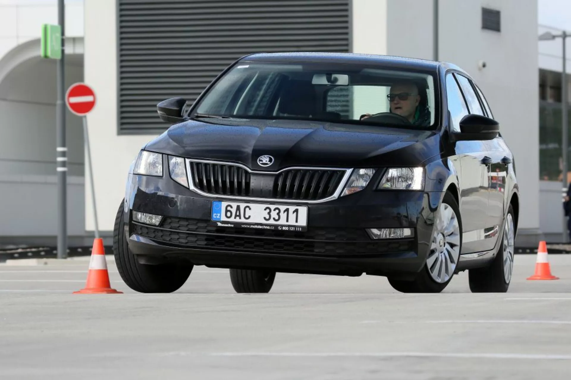 Czarna czterodrzwiowa sedan Hyundai i30 z 2017 roku nawigująca wokół pomarańczowych pachołków drogowych demonstrująca zdolności manewrowe pojazdu, widok z przodu i z boku z widocznym logo producenta na grillu oraz reflektory halogenowe lub ksenonowe bez wzorów LED, tradycyjna technologia wycieraczek, zamazane koła sugerujące ruch, i brak widocznych luksusowych akcesoriów lub zaawansowanych funkcji technologicznych, na utwardzonym placu testowym