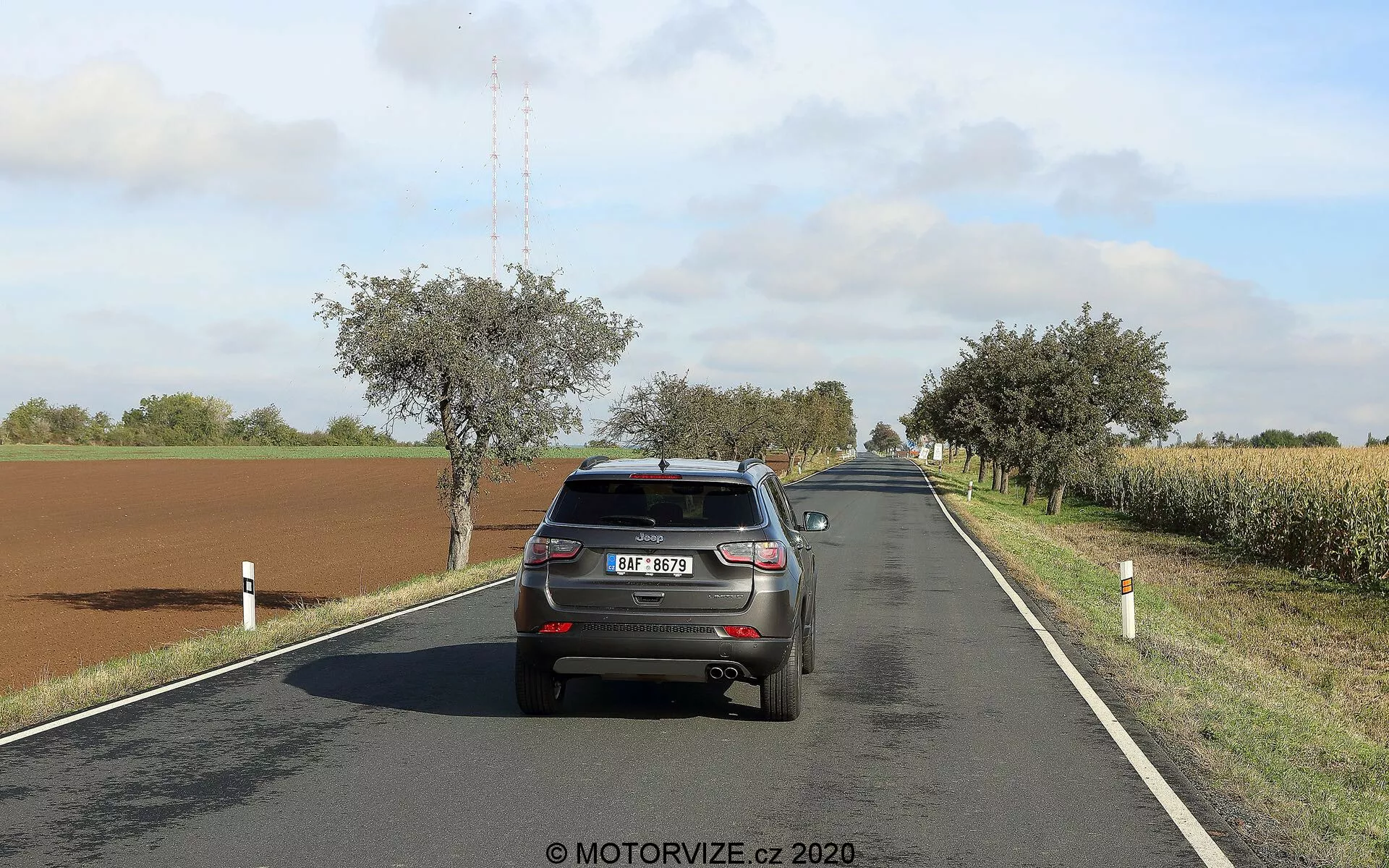 Obraz przedstawia tylny widok na Jeepa Compass z 2017 roku jadącego po dwupasmowej wiejskiej drodze w słoneczny dzień z rozproszonymi chmurami. Pojazd, znajdujący się po prawej stronie pasa, pokazuje swoją klapę tylną, zderzak, światła tylne, tylną szybę, tablicę rejestracyjną, część dachu z anteną w kształcie płetwy rekina oraz górną część tylnych opon. Samochód zajmuje znaczną część kadru, co pozwala na wyraźne zobaczenie jego cech. Droga łagodnie zakręca w prawo w oddali, otoczona polami, drzewami i przydrożnymi słupami. Światła hamowania samochodu nie są zapalone, co sugeruje normalną jazdę.