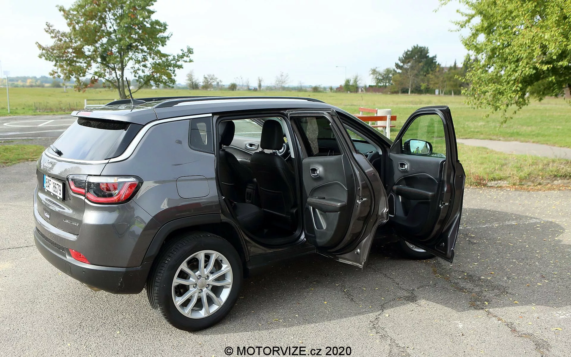 Obraz przedstawiający średniej wielkości SUV Jeep Compass 2017 zaparkowany na asfaltowej nawierzchni na terenie przypominającym park z trawą i drzewami w tle. Zdjęcie jest zrobione z kąta trzech czwartych od tyłu, ukazując profil i tylne części pojazdu. Obydwoje drzwi po stronie pasażera są otwarte, ujawniając ciemne materiałowe lub skórzane wnętrze, w tym przednie i tylne siedzenia, z możliwymi funkcjami takimi jak elektryczne okna i lusterka. SUV ma szare metaliczne wykończenie i jest wyposażony w pięcioramienne felgi aluminiowe, co może wskazywać na wyższy poziom wykończenia lub opcjonalne ulepszenie felg. Znak wodny 'MOTORVIZE.cz 2020' sugeruje, że obraz pochodzi z recenzji samochodu lub strony internetowej o tematyce motoryzacyjnej.