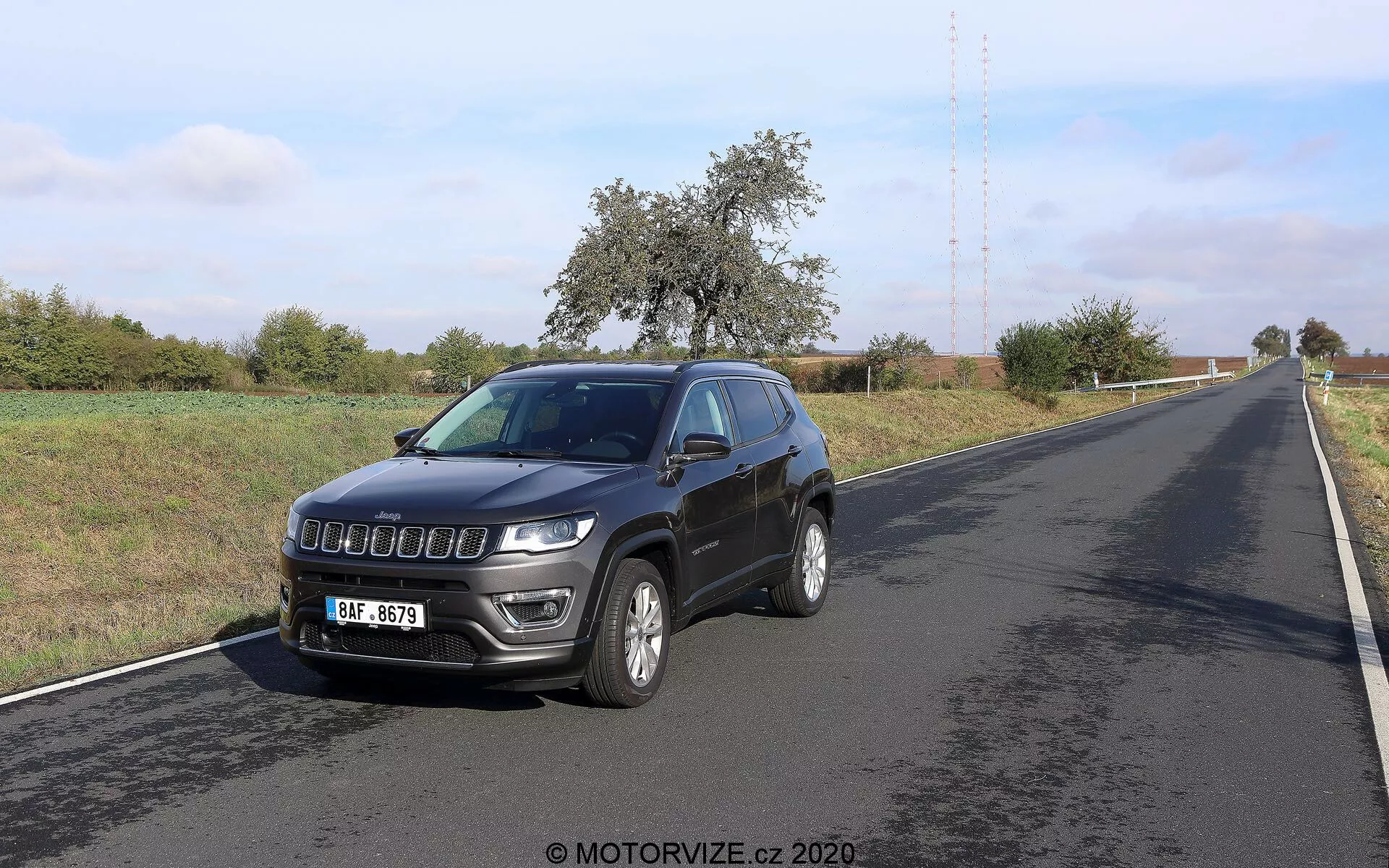 Zdjęcie Jeep Compass 2017 sfotografowanego z przedniego trójkątnego kąta. Pojazd, umieszczony po prawej stronie dwupasmowej drogi, pokazuje swój przedni i boczny profil. Kluczowe cechy obejmują nowoczesną kratkę, wyraźne reflektory z diodami LED do jazdy dziennej, aluminiowe felgi, zintegrowane kierunkowskazy na lusterkach bocznych, antenę dachową i światła przeciwmgielne na przednim zderzaku. Otoczenie wydaje się wiejskie, z czystym niebem i roślinnością wzdłuż drogi.