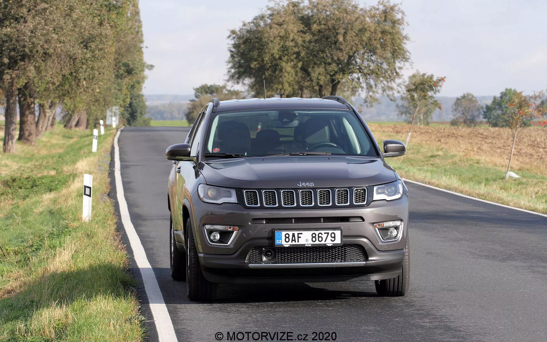 Obraz Jeep Compass 2017 jadącego dwupasmową drogą, sfotografowanego z przedniego trójwymiarowego kąta. Ikoniczna, siedmioszczelinowa krata pojazdu, szczegółowe reflektory ze światłami do jazdy dziennej LED, światła przeciwmgielne i wieloszprychowe aluminiowe koła są wyraźnie widoczne. Szary lub czarny kolor Jeepa nadaje mu solidny i solidny wygląd, a naturalne światło dzienne podkreśla jego cechy. Pojazd wydaje się być w ruchu, z lekkim rozmyciem kół. Drzewa i otwarte pola w tle sugerują wiejskie lub półwiejskie otoczenie. Samochód zajmuje środkową i dolną połowę kadru, nieco z prawej strony, ukazując przednią kratę, reflektory, zderzak, maskę, błotnik po stronie kierowcy, koło i przednie drzwi.