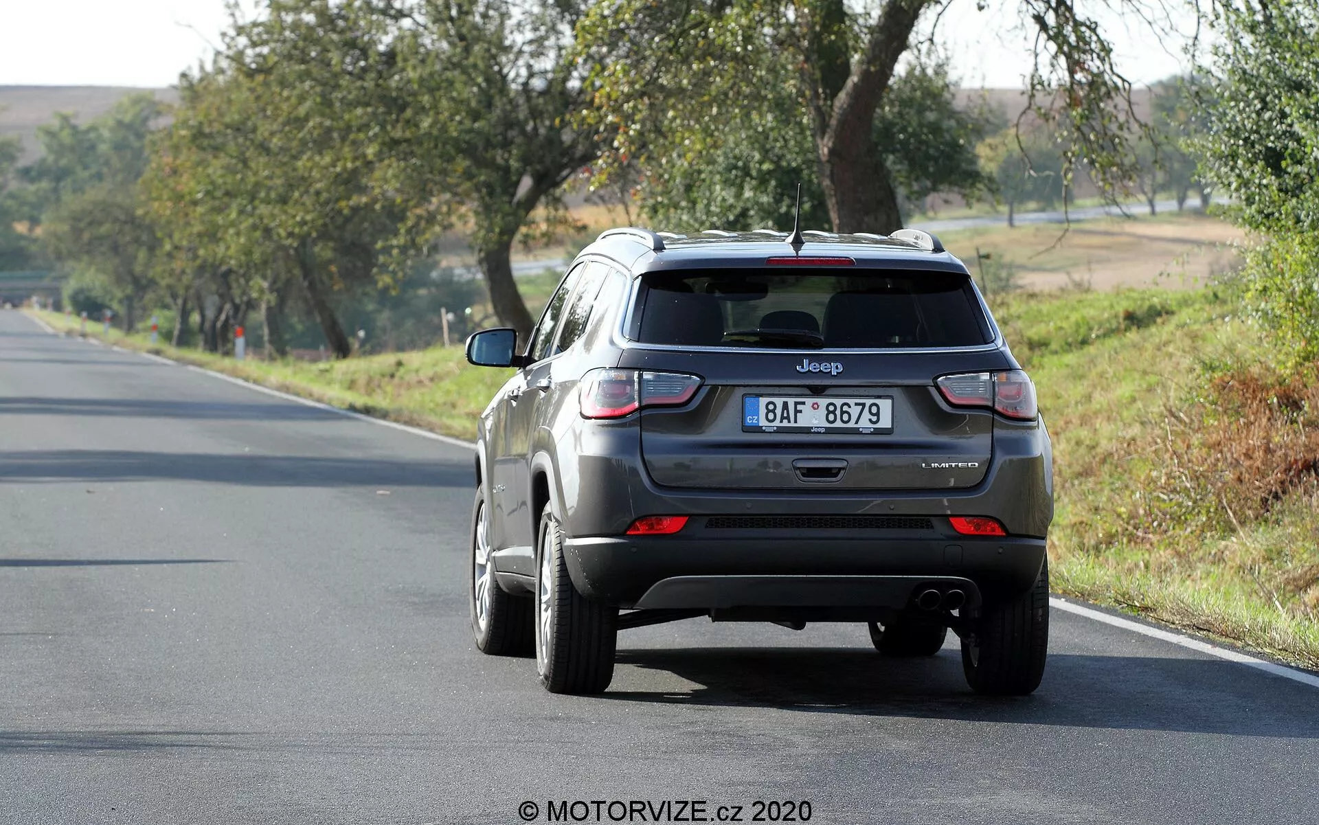Widok z tyłu metalicznego szarego SUV-a Jeep Compass z 2017 roku, jadącego w oddali po dwupasmowej drodze, umieszczonego nieco po prawej stronie kadru. Pojazd, na prawym pasie, posiada widoczne cechy, takie jak tylna klapa z emblematem Jeep i tablicą rejestracyjną, tylna szyba z małym spojlerem, tylne światła z czerwonymi i białymi sekcjami, tylny zderzak z czujnikami oraz antenę w kształcie płetwy rekina na dachu. Felgi aluminiowe są częściowo widoczne. Scena sugeruje słoneczny dzień z okolicznymi drzewami i trawą, co wskazuje na wiejskie lub półwiejskie otoczenie.