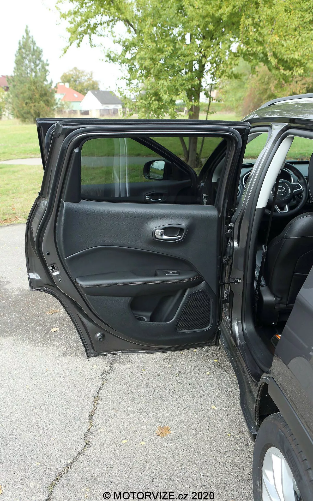 Widok wnętrza Jeep Compass 2017 z zewnątrz pokazujący otwarte drzwi kierowcy z metalową i ergonomiczną klamką, schowkami, kratką głośnika i panelem sterowania do obsługi okien i zamków drzwi. Widoczna jest część deski rozdzielczej, konsoli centralnej i wielofunkcyjnej kierownicy, co sugeruje nowoczesne, bogate wnętrze. Światło naturalne, pochmurna pogoda, drzewa w tle sugerujące zewnętrzne otoczenie w parku lub na ulicy wysadzanej drzewami.