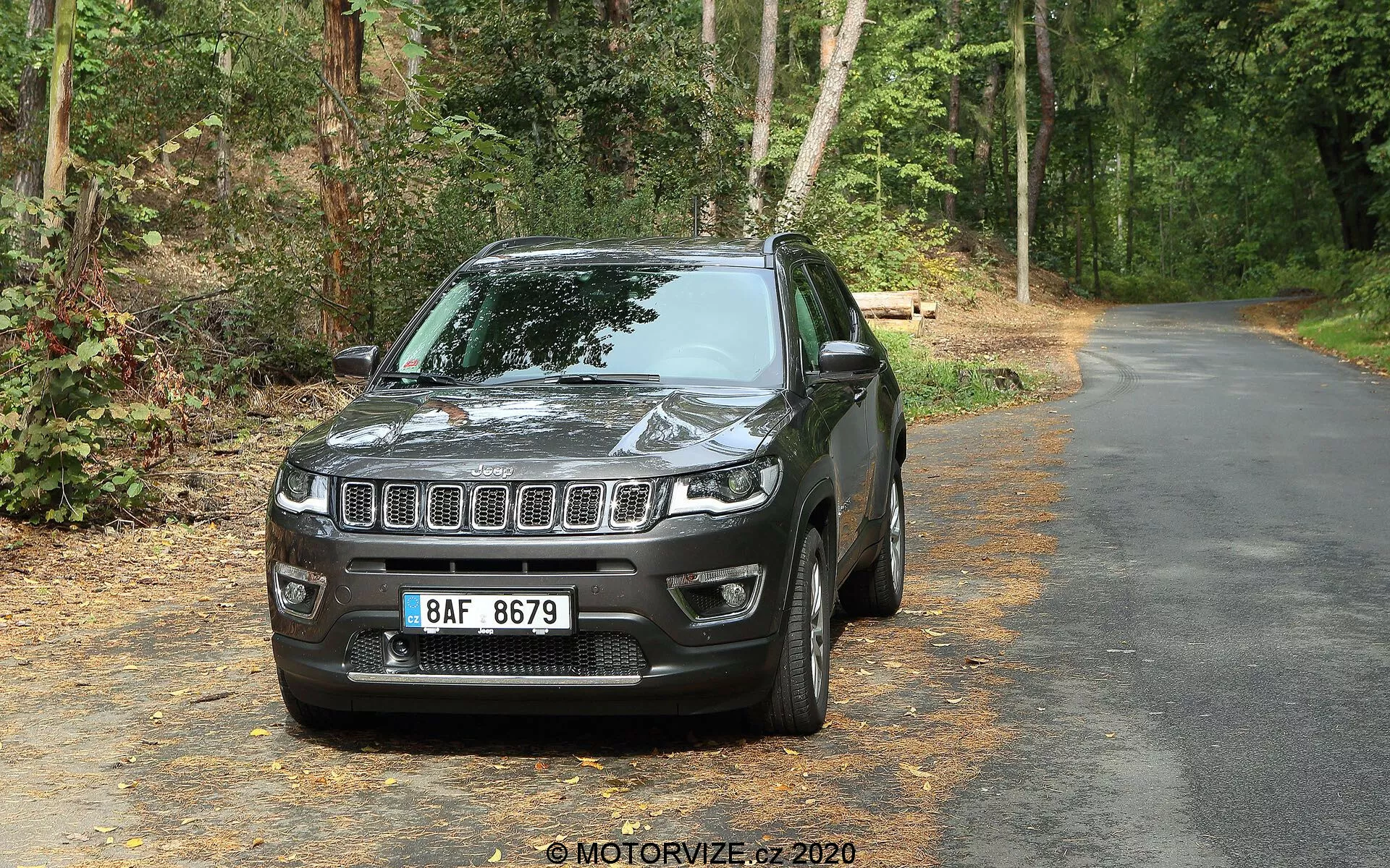 SUV Jeep Compass 2017 na utwardzonej drodze otoczonej zielenią, ustawiony przodem do kamery pod lekkim kątem w lewo, ukazując przód i stronę kierowcy. Widoczne cechy to pozioma atrapa chłodnicy z centralnym emblematem, prostokątne reflektory LED, obudowy świateł przeciwmgielnych w dolnym zderzaku, konturowana maska odbijająca światło, lusterka boczne w kolorze nadwozia, lekko przyciemniane szyby, wieloramienne felgi aluminiowe i wyraźnie zarysowany błotnik. W tle drzewa i liście sugerują obszar leśny przy drodze.