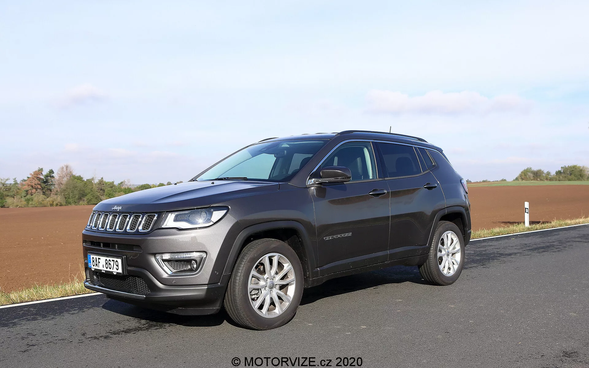 Nowoczesne SUV zaparkowane przy drodze z polem w tle, uchwycone z kąta przedniego trzy czwarte. Zdjęcie ukazuje siedmioszczelinowy grill przedni SUV-a, charakterystyczne reflektory wyposażone w diody LED, felgi aluminiowe i ciemny metaliczny lakier. Widoczne są szczegóły boczne, takie jak linia okien, klamki drzwi, lusterka boczne prawdopodobnie z kierunkowskazami i lekko opadająca linia dachu. Na przednich drzwiach widoczna jest także odznaka, która może dostarczyć informacji o wersji wyposażenia.