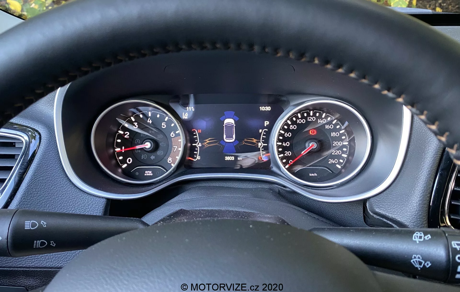 Zbliżenie na panel instrumentów Jeep Compass 2017 z perspektywy kierowcy, pokazujące zarówno wskaźniki analogowe, jak i centralny wyświetlacz cyfrowy. Funkcje obejmują obrotomierz, prędkościomierz z podwójną jednostką, wskaźnik wyboru biegu, wskaźnik paliwa, licznik kilometrów oraz dodatkowe wskaźniki technologii bezpieczeństwa, takie jak asystent pasa ruchu. Szwy na kierownicy z materiału podobnego do skóry podkreślają jakość i komfort, a dźwignie i przyciski sugerują zaawansowane funkcje wygody i asystenta kierowcy.