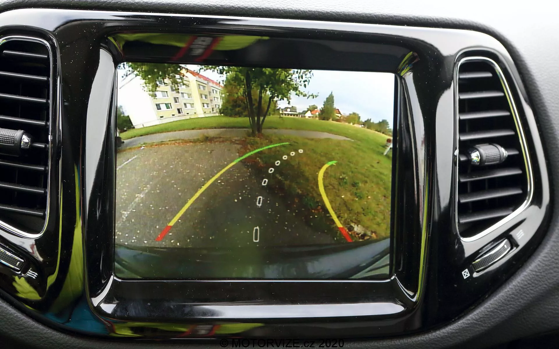Obraz systemu kamery cofania na ekranie deski rozdzielczej w samochodzie Jeep Compass 2017. Kamera, umieszczona z tyłu pojazdu w pobliżu tablicy rejestracyjnej, zapewnia szerokokątny widok za samochodem. Widoczne są linie pomocnicze do cofania, wskazujące szerokość pojazdu i przewidywany tor jazdy za pomocą żółtych i czerwonych linii określających odległość. Scena przedstawia parking lub podjazd z drzewami i trawiastym obszarem w tle. Ekran jest umieszczony centralnie na desce rozdzielczej, otoczony otworami wentylacyjnymi klimatyzacji, co świadczy o wysokiej jakości obrazu, typowej dla nowoczesnych technologii samochodowych.
