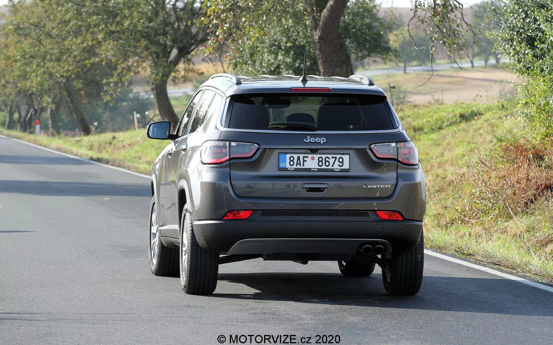 Widok tylnej części srebrnego Jeepa Compass 2017, ukazujący pokrywę bagażnika, tylny zderzak i światła tylne. Widoczny jest napis 'Jeep' oraz nazwa modelu 'Compass', z lekko nieczytelną tabliczką oznaczającą poziom wykończenia. Obraz wyraźnie pokazuje światła tylne, końcówkę wydechu pod zderzakiem i samochód ustawiony na dwupasmowej drodze otoczonej drzewami w wiejskiej scenerii.