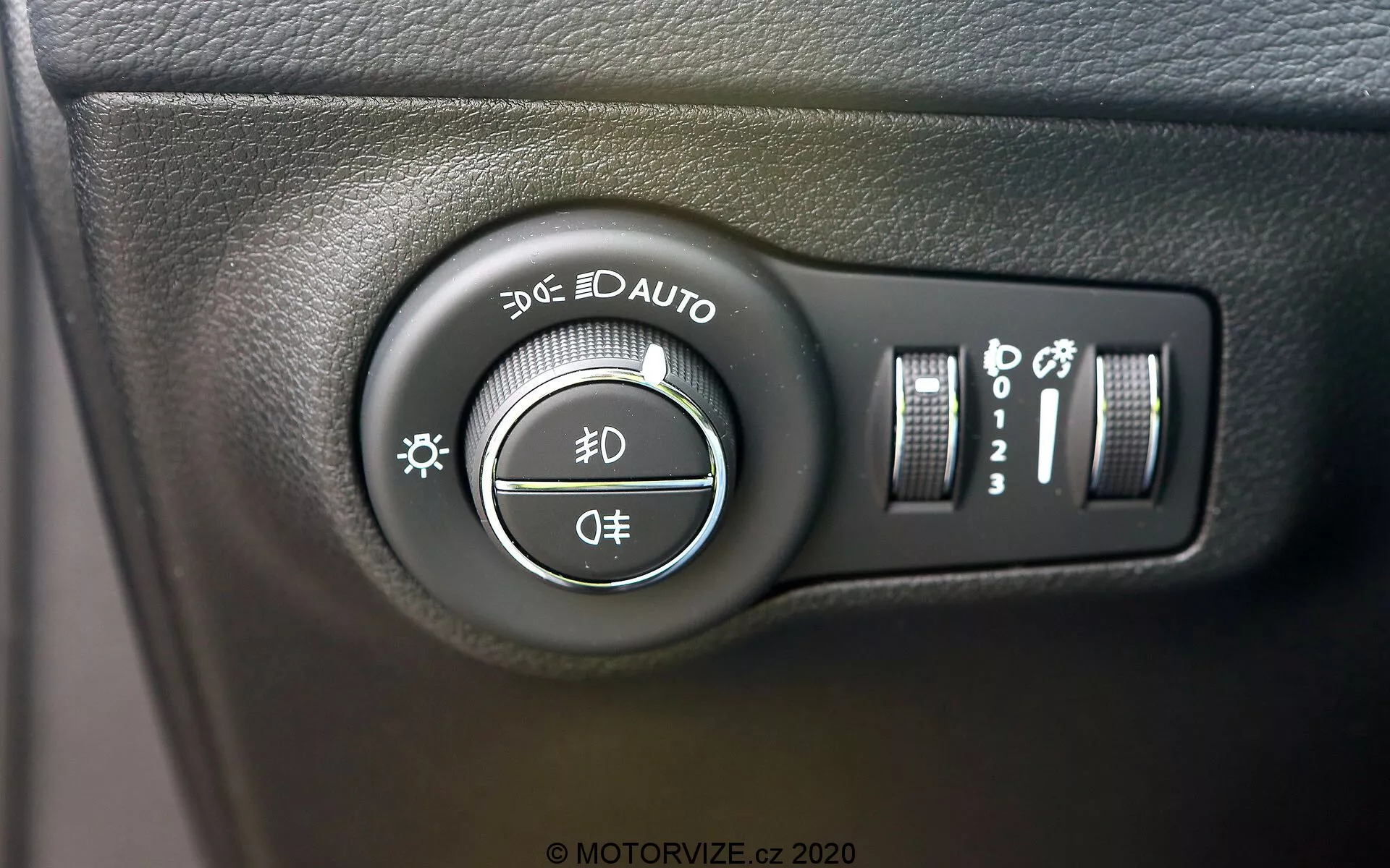 Zbliżenie przełącznika świateł Jeep Compass 2017 umieszczonego na desce rozdzielczej po lewej stronie kierownicy. Zdjęcie wykonane pod niewielkim kątem podkreśla pokrętło z ustawieniami do świateł postojowych, świateł do jazdy dziennej, automatycznych świateł, świateł mijania i świateł drogowych oraz pozycji wyłączenia. Dodatkowe kontrolki po prawej stronie to pionowy suwak do regulacji jasności deski rozdzielczej i przycisk, który może wskazywać automatyczne światła drogowe lub poziomowanie świateł. Przełącznik zamontowany jest na powierzchni z fakturowanego czarnego plastiku, z wyraźnymi białymi oznaczeniami i lekkimi, błyszczącymi refleksami.