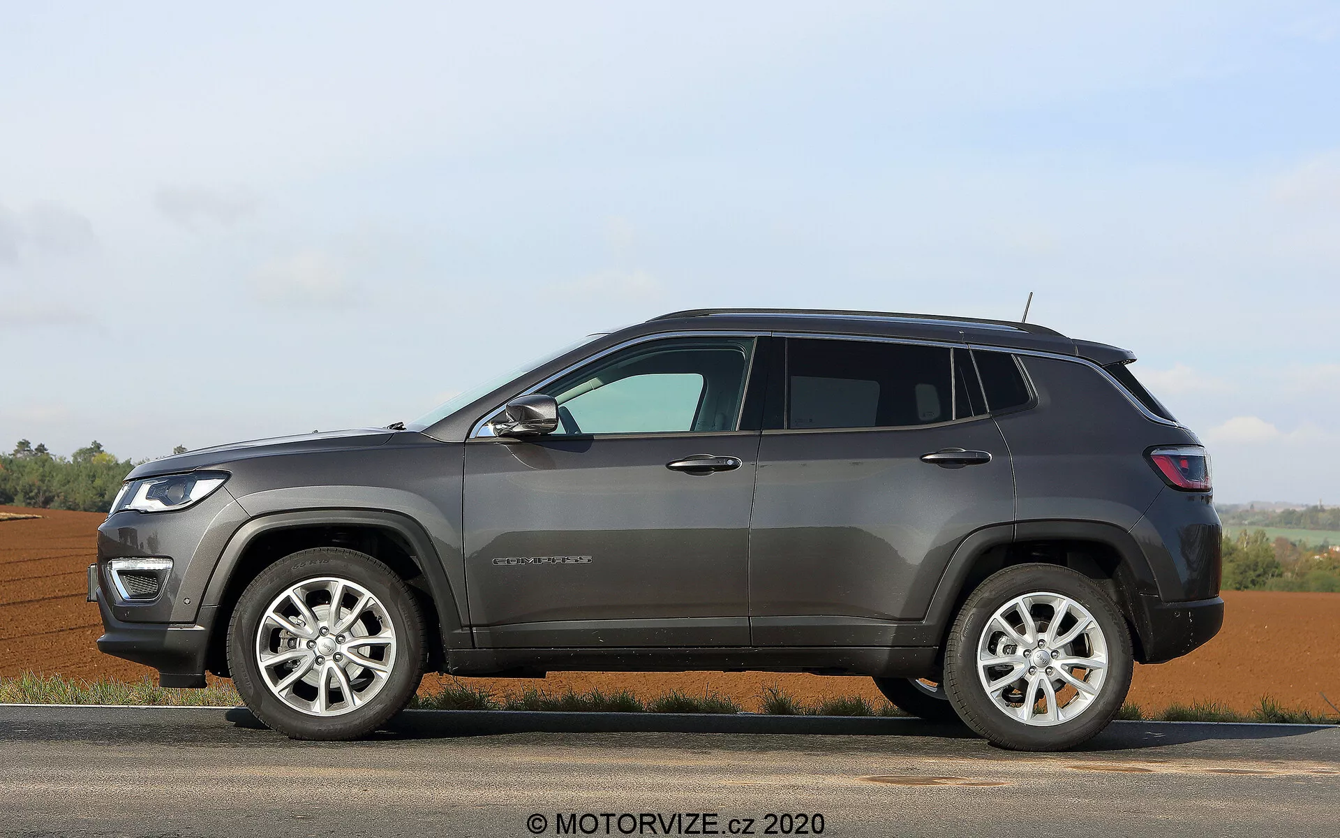 Widok z profilu bocznego nowoczesnego SUV-a Jeep Compass 2017 zaparkowanego na równej powierzchni, przedstawiający jego prawą stronę z podniesionymi szybami, czterema drzwiami, widocznym słupkiem B i okienkiem ćwiartkowym w rejonie słupka C. Widoczne są aluminiowe felgi z wieloramiennym wzorem i opony z wyraźnym bokiem, wskazujące na możliwość użytkowania zarówno na drodze, jak i poza nią. Zewnętrzne cechy obejmują plastikowe nakładki wokół nadkoli i dolnej części nadwozia, relingi dachowe do dodatkowego ładunku, boczne lusterka prawdopodobnie z wbudowanymi kierunkowskazami oraz wyraziste kontury drzwi. Na przednim błotniku znajduje się emblemat wskazujący model lub poziom wykończenia, a podwyższony prześwit pojazdu sugeruje wysoką prześwity, odpowiednie do różnych terenów. Stacjonarny, na tle wiejskim z otwartymi polami i czystym horyzontem, w łagodnych warunkach oświetleniowych.