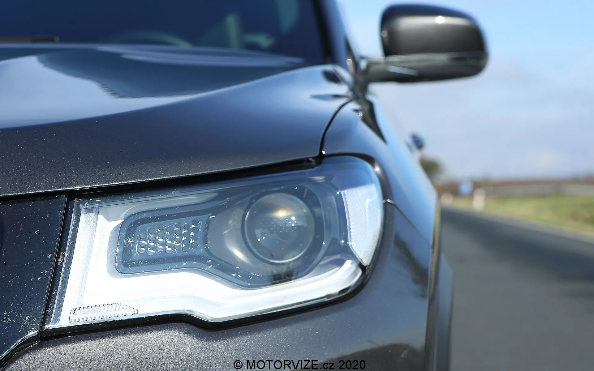 Zbliżenie lewej przedniej strony Jeep Compass 2017, koncentrujące się na zestawie reflektora w niskim kącie przy poziomie gruntu. Nowoczesny reflektor zawiera projektor oraz światła do jazdy dziennej LED rozmieszczone we wzór charakterystyczny. Obudowa reflektora, elegancka, gładko integruje się z ciemną karoserią, odbijając jasne niebo. Boczny lusterko, częściowo widoczne, pasuje do koloru nadwozia i może zawierać boczny wskaźnik. Płytka głębia ostrości podkreśla szczegóły reflektora i przednią kratkę na rozmytym tle drogi.
