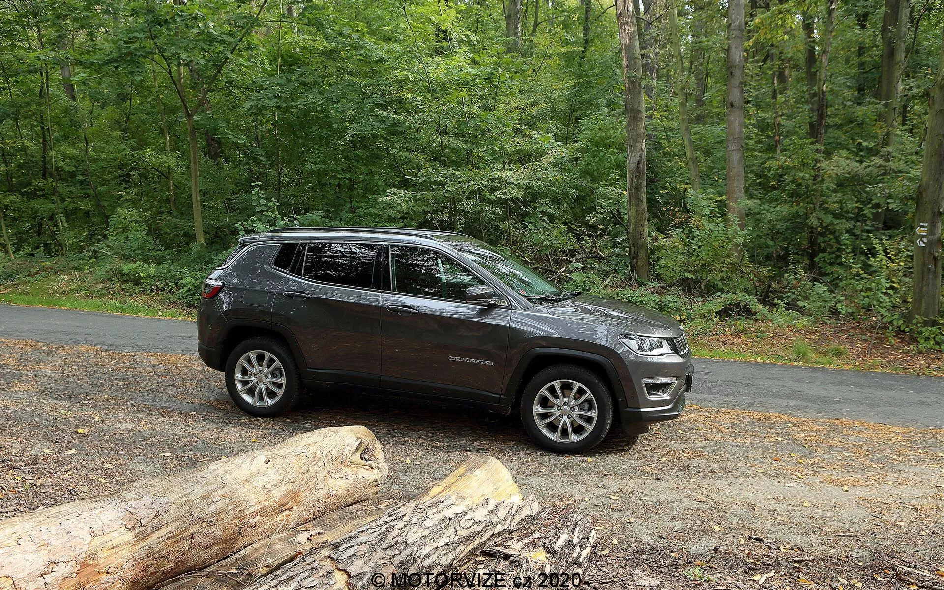 Widok z boku na SUV Jeep Compass 2017 zaparkowany na asfaltowej drodze w naturalnym otoczeniu z bujnymi zielonymi drzewami. Pojazd pokazuje nowoczesne linie projektowe wzdłuż drzwi i nadkoli, z widocznymi felgami ze stopu o wieloszprychowym wzorze. Ma wysoki prześwit, co sugeruje możliwości terenowe. Zdjęcie uchwyca część przedniego grilla i reflektorów, wskazując na współczesny styl. Widoczne są relingi dachowe, zintegrowane kierunkowskazy w lusterkach bocznych i przyciemniane szyby. Ciemny lakier z błyszczącym wykończeniem wskazuje na dobrze utrzymany pojazd. Stacjonarny SUV widoczny jest przy obfitym naturalnym świetle, idealnie nadający się do pokazania jego cech i atrakcyjności dla entuzjastów outdoorowych przygód.