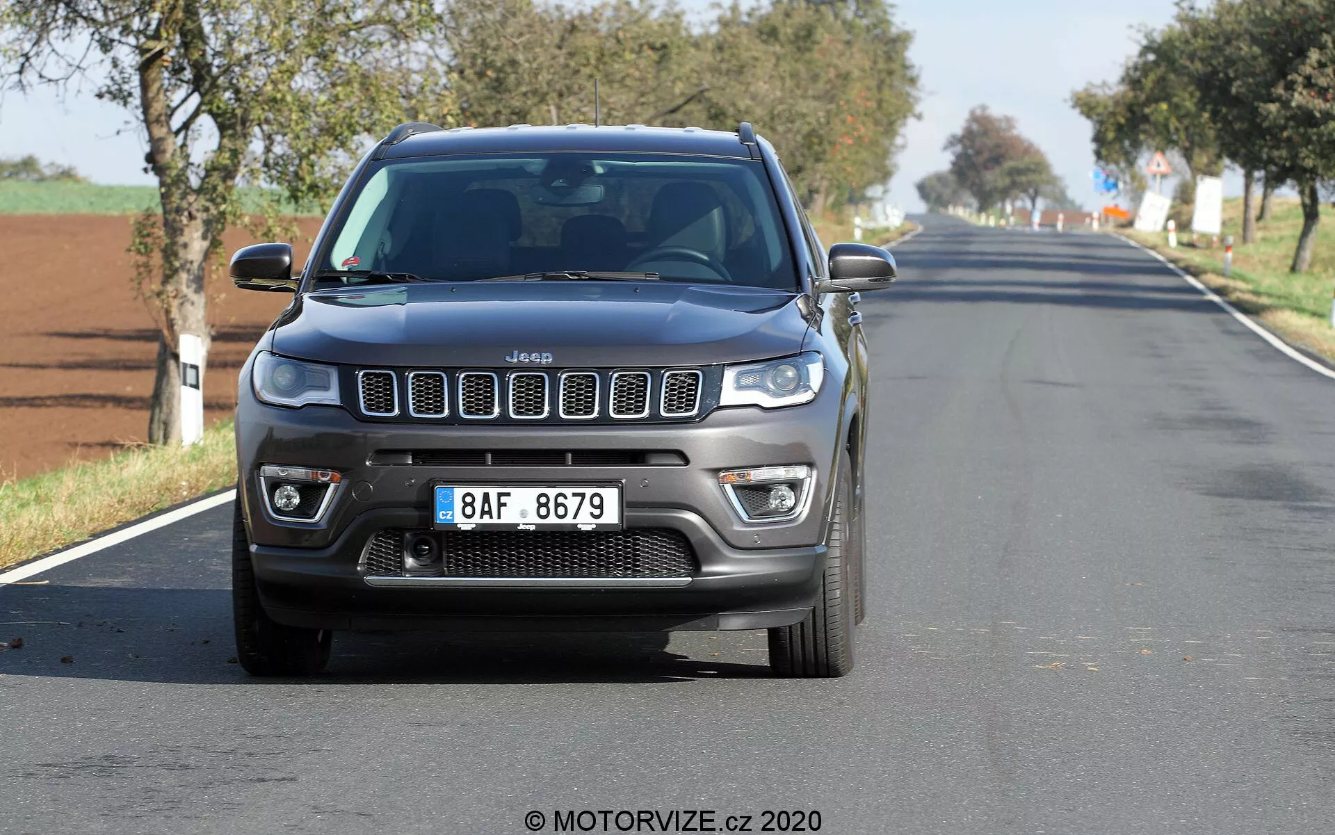 Widok z przodu Jeep Compass 2017 jadącego dwupasmową, wiejską lub podmiejską drogą, uchwycony pod niskim kątem, ukazujący ikoniczny grill z siedmioma szczelinami, LED-owe światła do jazdy dziennej, światła przeciwmgielne i wyraziste kontury maski. Samochód jest wyśrodkowany i stosunkowo blisko kamery, z lekkim rozmyciem kół i tła. Czyste niebo sugeruje jasny, słoneczny dzień.