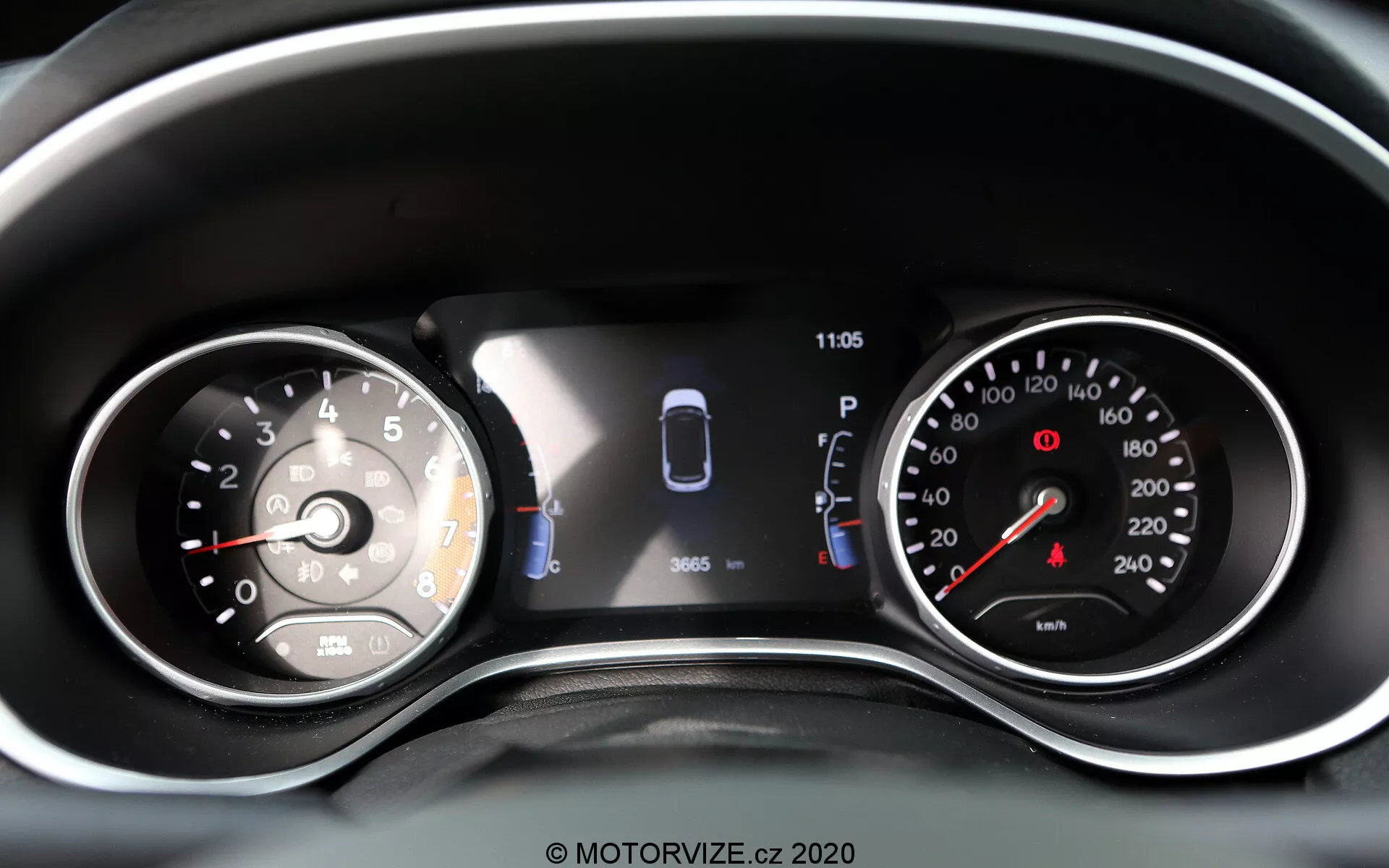 Zbliżenie na zestaw wskaźników Jeep Compass 2017 z kombinacją analogowych tarcz i wyświetlacza cyfrowego. Po lewej stronie znajduje się analogowy prędkościomierz oznaczony do 260 z igłą wskazującą nieco powyżej 0. Centralny wyświetlacz cyfrowy pokazuje grafikę auta z czujnikami oraz dwiema otwartymi drzwiami, wskazując asystenta parkowania i status pojazdu. Zegar pokazuje godzinę 11:05. Po prawej stronie znajduje się analogowy obrotomierz z czerwonym polem zaczynającym się przed 7, oznaczony do 8 (przyrosty 1,000 obr./min). Widoczne są dodatkowe kontrolki ostrzegawcze i ikony, co czyni to dobrze oświetloną i czytelną reprezentacją nowoczesnych i tradycyjnych odczytów pojazdu oraz technologii wspomagania kierowcy.