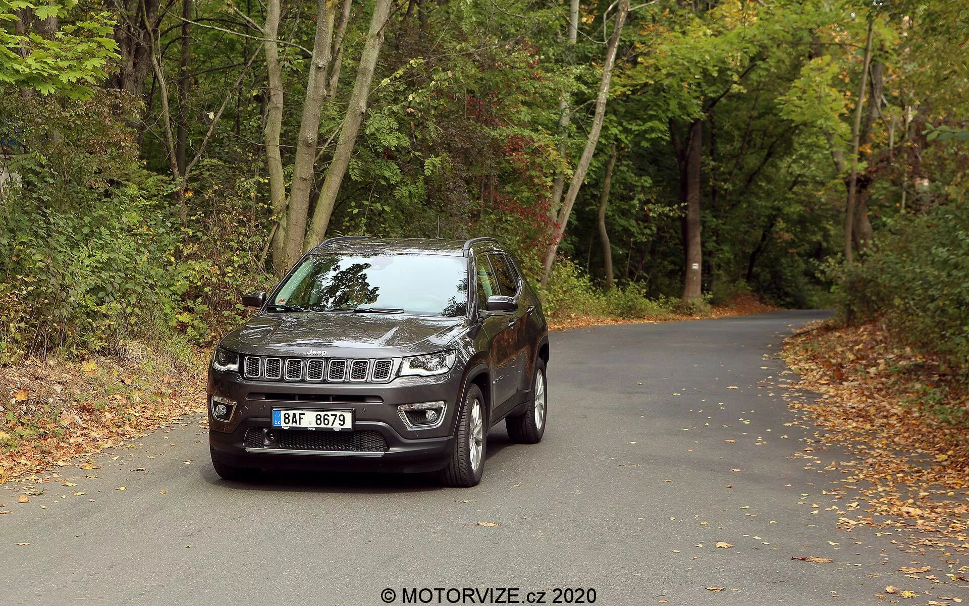 SUV Jeep Compass 2017 ustawiony na asfaltowej drodze w otoczeniu lasów z jesiennymi liśćmi. Pojazd sfotografowano z przedniego, trzewokrotnego kąta, ukazując charakterystyczny grill Jeepa z siedmioma szczelinami i centralnym emblematem marki, nowoczesne reflektory LED oraz światła przeciwmgielne wmontowane w zderzak. Jest wyposażony w felgi aluminiowe, lusterka boczne z wbudowanymi kierunkowskazami, solidną konstrukcję z obudową nadkoli i lekko przyciemniane szyby. Szczegółowy widok podkreśla elementy designu i technologii, sugerując jego przydatność zarówno na miejskich, jak i terenowych przygodach.