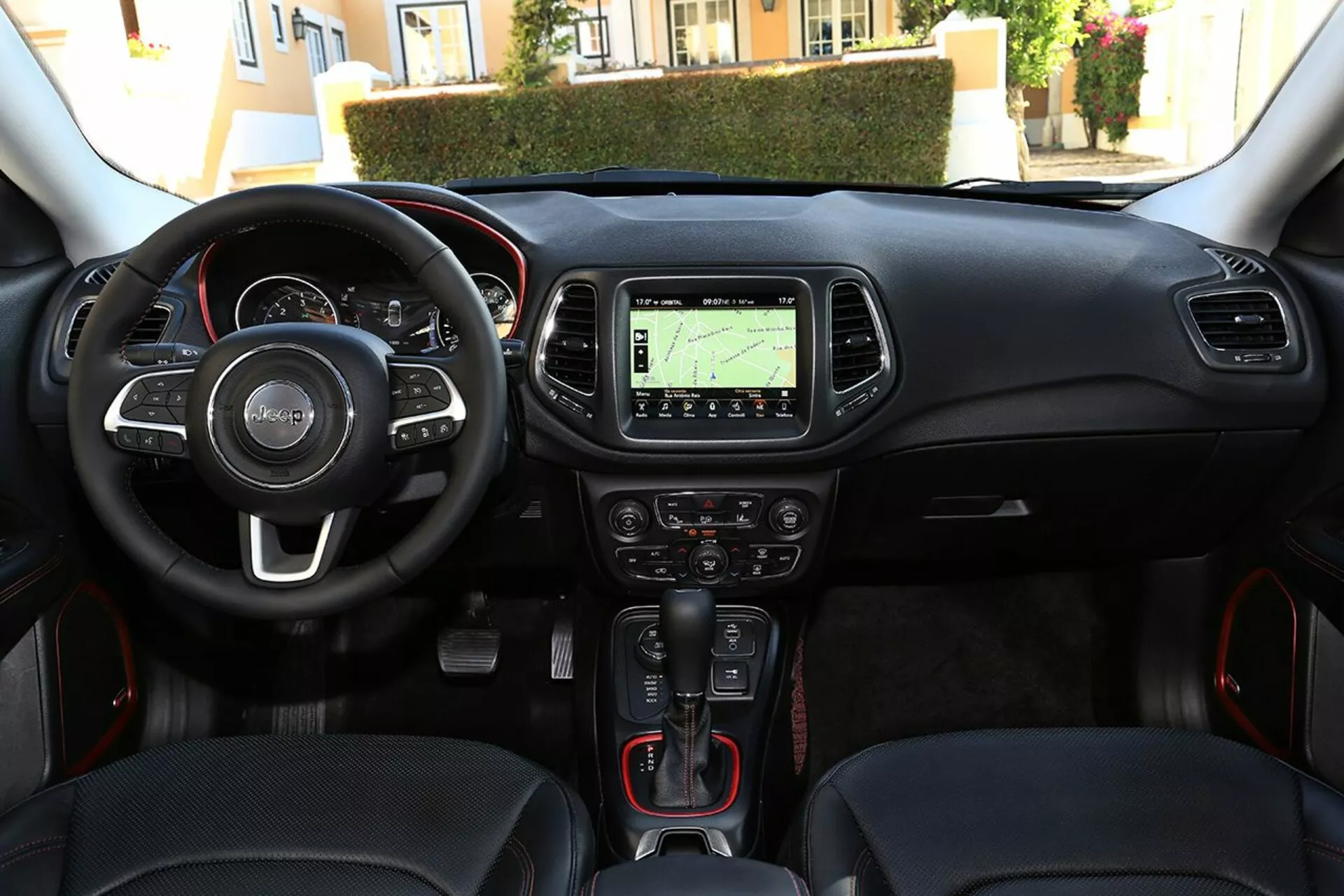 Widok wnętrza Jeep Compass 2017 z perspektywy kierowcy, pokazujący kierownicę z zamontowanymi kontrolami, zespół wskaźników, konsolę środkową z systemem nawigacji, kontrole klimatyzacji, automatyczną dźwignię zmiany biegów z sportowymi czerwonymi detalami, guzik uruchamiania-zatrzymywania bez kluczyka i port łączności multimedialnej