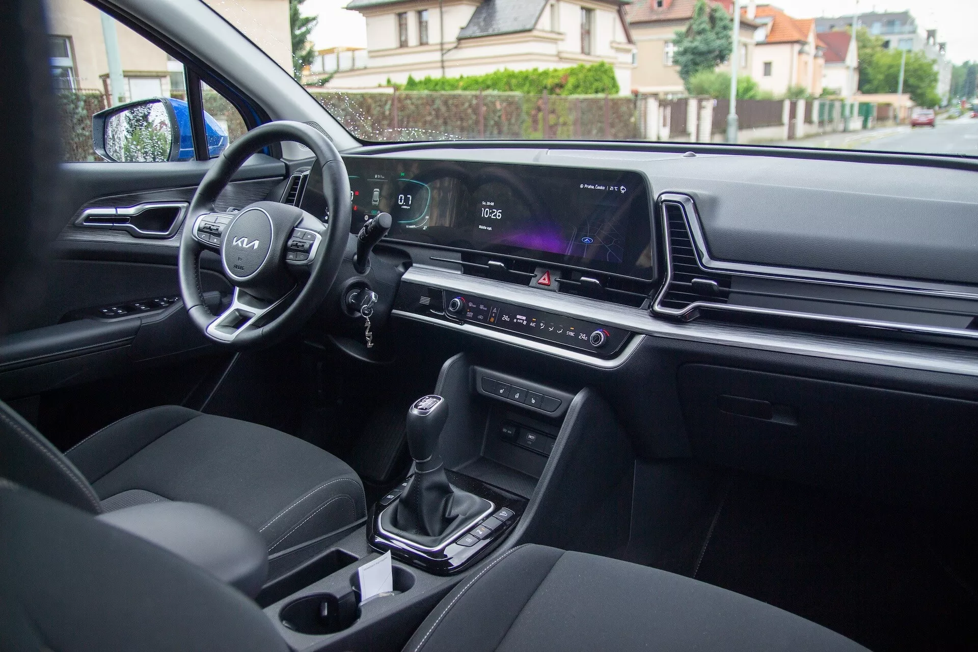 Wnętrze Kia Sportage 2022 widziane z tylnego siedzenia, pokazujące sterowanie na drzwiach kierowcy, kierownicę z logo i zamontowane sterowania, cyfrowy zestaw wskaźników, ekran systemu multimedialnego, kontrolę klimatyzacji, automatyczną dźwignię zmiany biegów, uchwyty na kubki i nowocześnie tapicerowane fotele