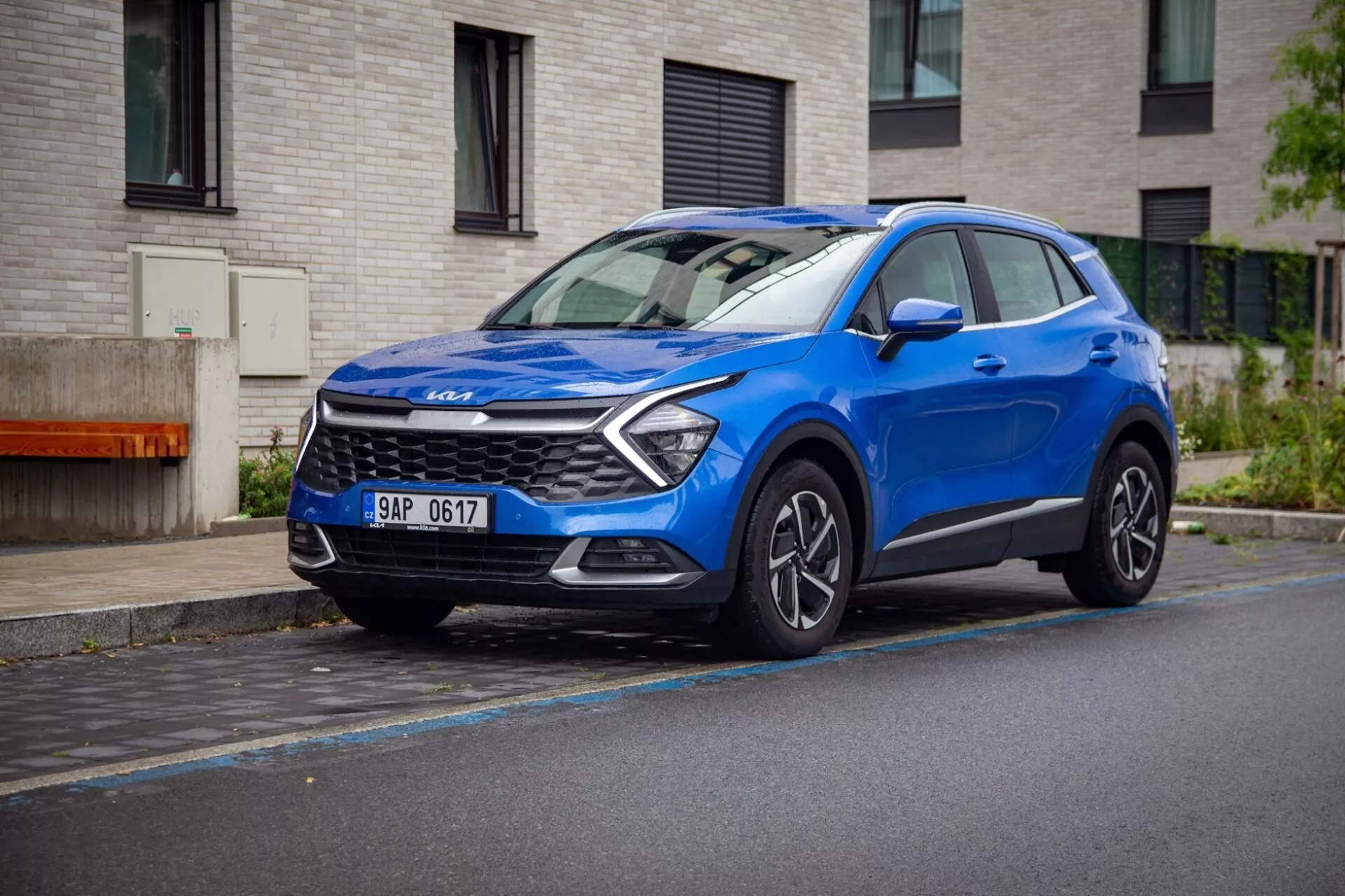 Kompaktowy SUV crossover Kia Sportage 2022 w kolorze niebieskim, zaparkowany na mokrej ulicy, prezentujący dynamiczny trzy czwarte widok z przodu z diodami LED, rzeźbioną maską i felgami ze stopu.