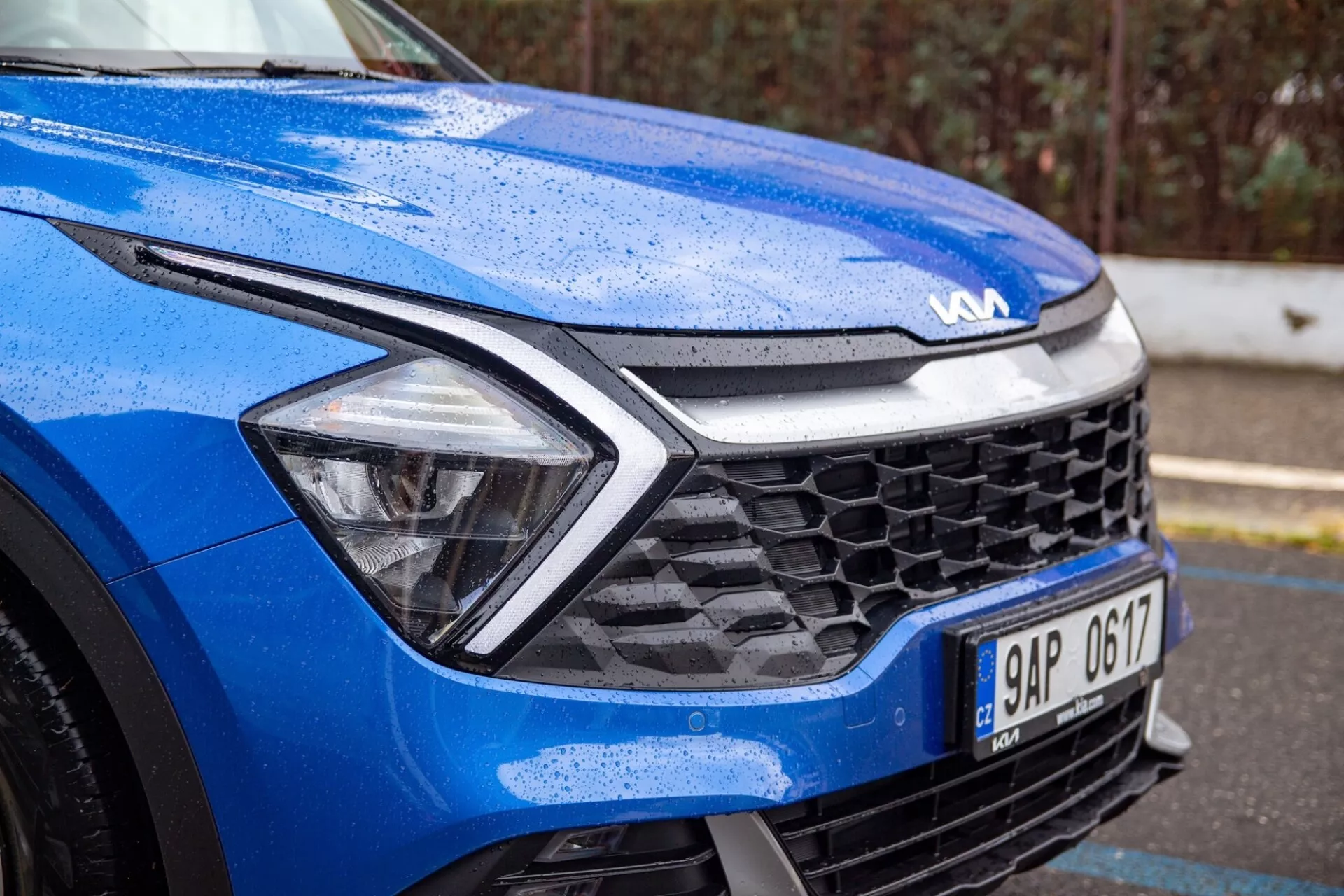 Zbliżony widok na niebieskiego Kia Sportage 2022 skupiony na przedniej lewej ćwiartce w wilgotnych warunkach, podkreślający dynamiczny design maski, reflektory LED, grilla plaster miodu oraz zaawansowaną technologię sensorów