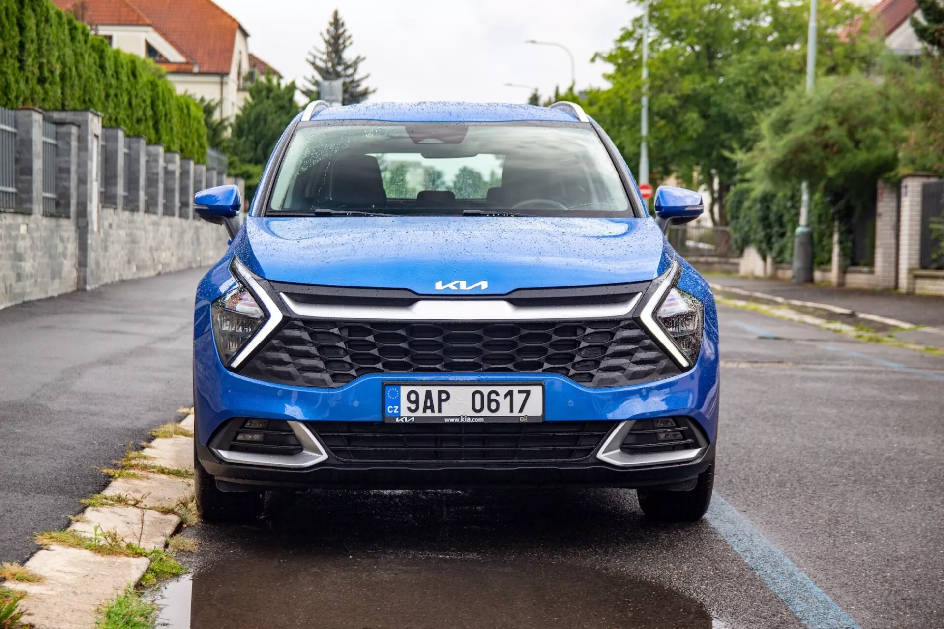 Widok z przodu na niebieskiego Kia Sportage z 2022 roku, zaparkowanego na przedmieściach, prezentującego nowoczesną kratę, eleganckie reflektory, chromowane logo Kia i dynamiczny design przedniej części