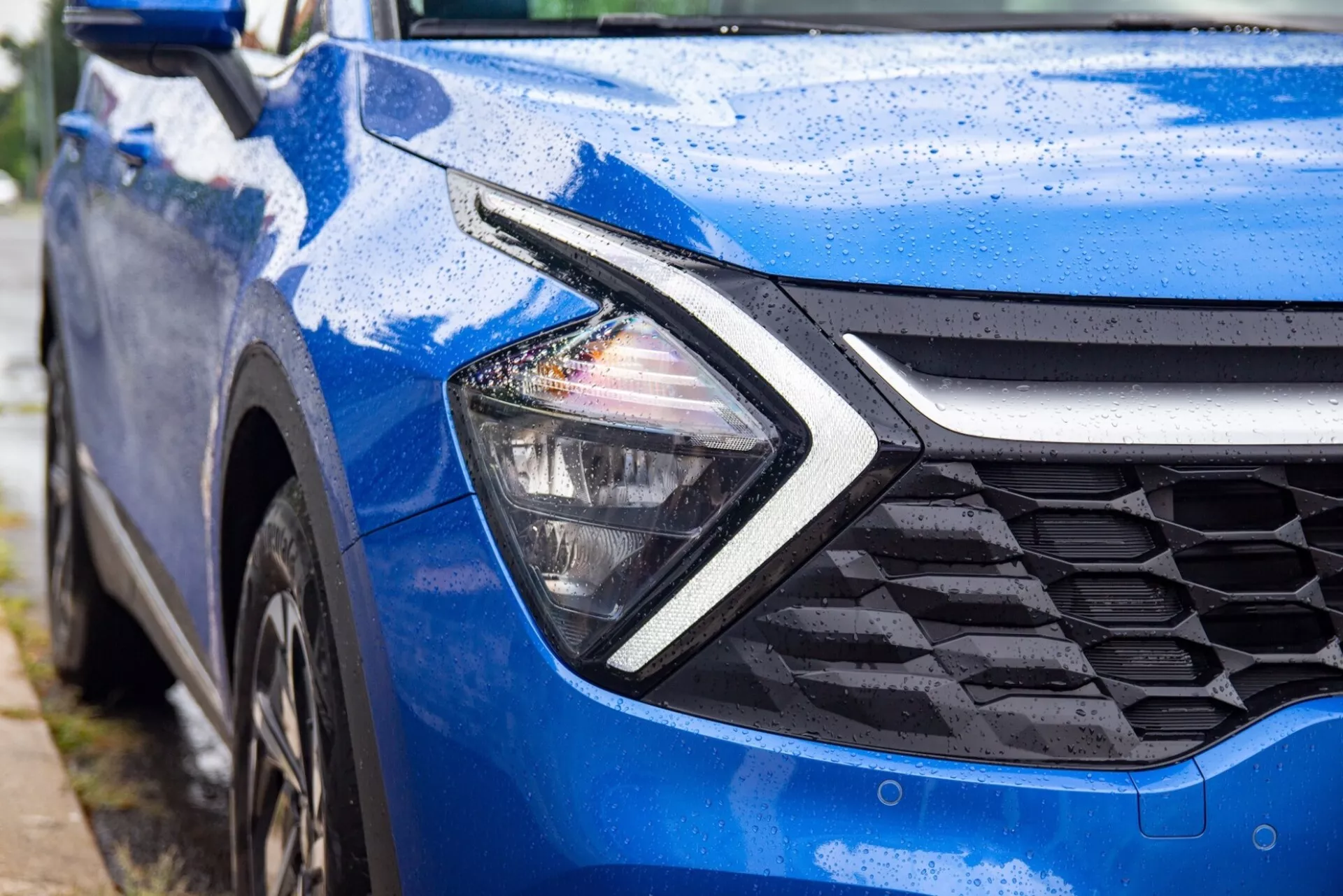 Kia Sportage 2022 w kolorze niebieskim, prezentująca nowoczesne reflektory LED z charakterystycznym podświetleniem w kształcie bumerangu i wzorzystą kratę z perspektywy czołowo-bocznej, podkreśloną kroplami wody