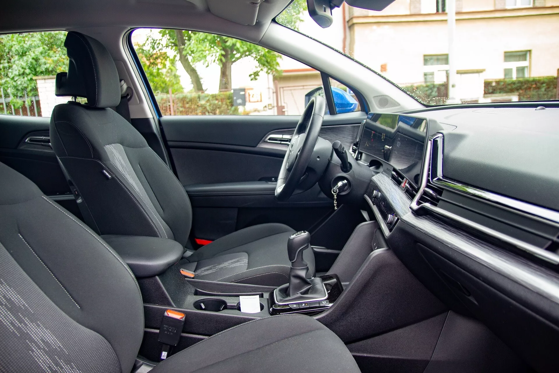 Widok wewnętrzny Kia Sportage 2022 z perspektywy tylnego pasażera przedstawiający przednie siedzenie pasażera, konsolę centralną z automatyczną dźwignią zmiany biegów, kierownicę po lewej stronie i duży ekran systemu infotainment na desce rozdzielczej