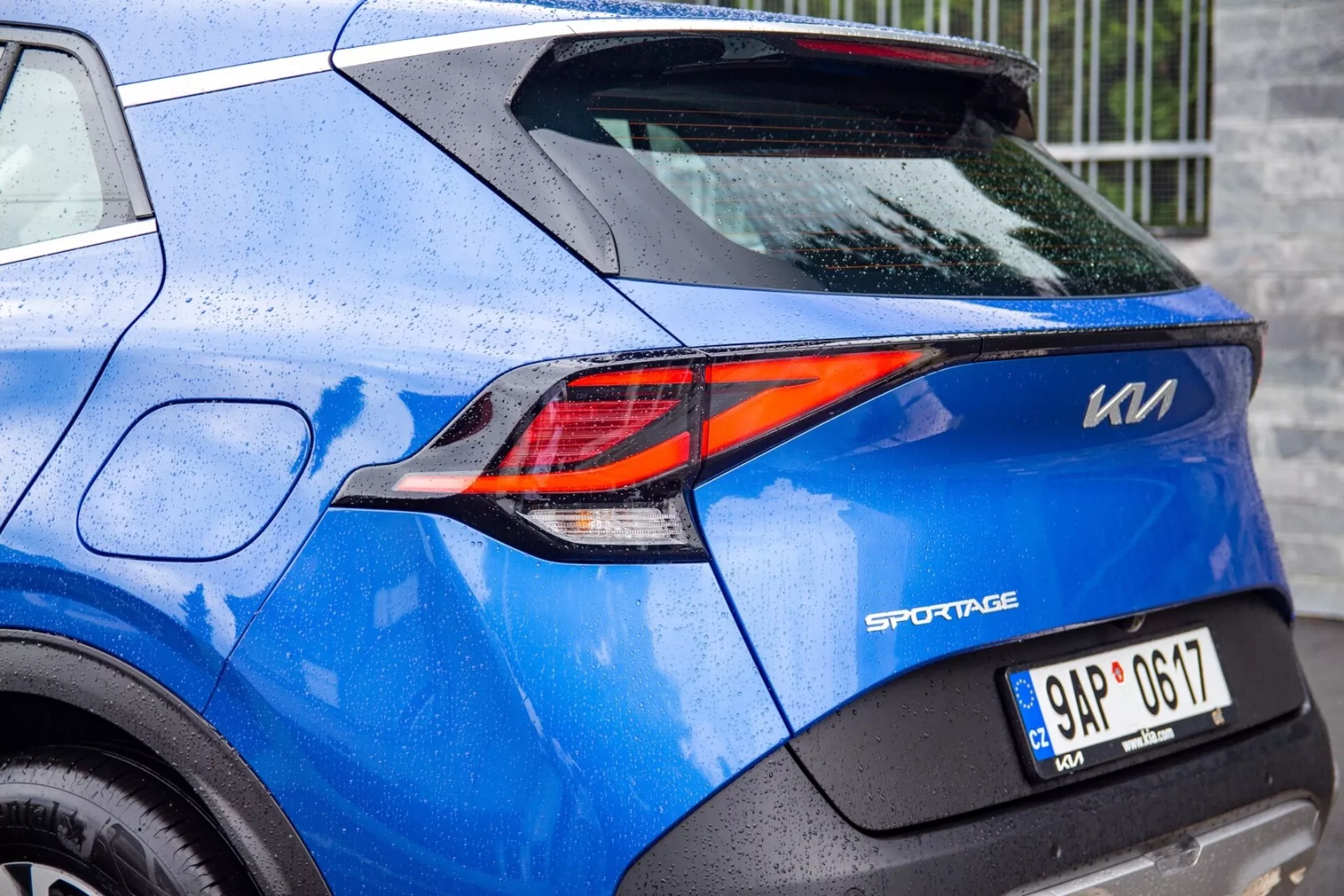 Częściowy widok niebieskiego Kia Sportage 2022 podkreślający wyszukane tylne światło LED w kształcie litery Z i szczegóły konstrukcji tylnej, w tym odznaka modelu, tablica rejestracyjna i lakier pokryty kroplami deszczu