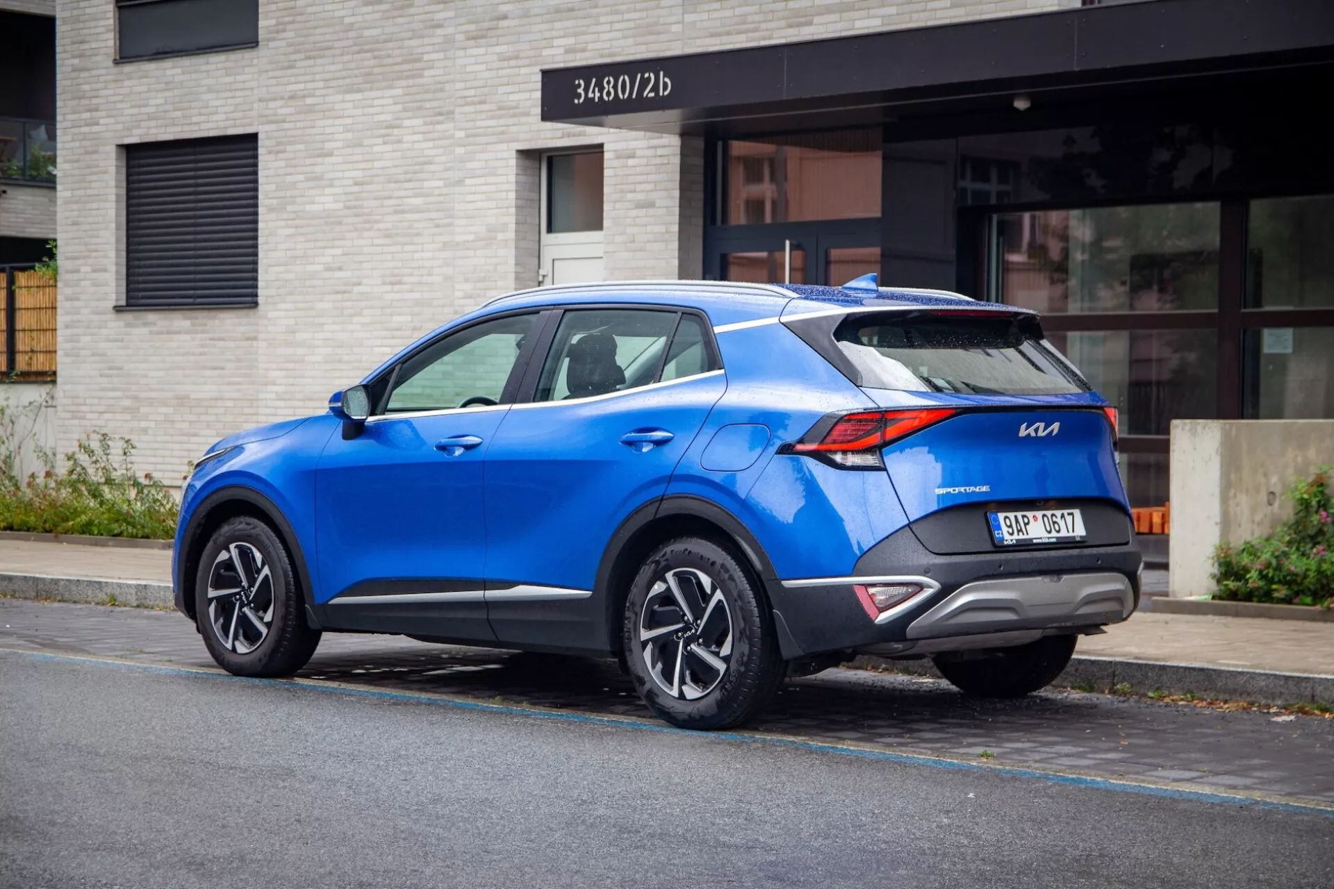 Nowoczesne SUV crossover Kia Sportage 2022 zaparkowane na ulicy miejskiej, prezentujące widok z tyłu trzy czwarte z wyraźnymi światłami tylnymi, sportowym spojlerem na dachu, aluminiowymi felgami wieloramienne, żywym niebieskim kolorem oraz potencjalnymi nowoczesnymi funkcjami technologicznymi.