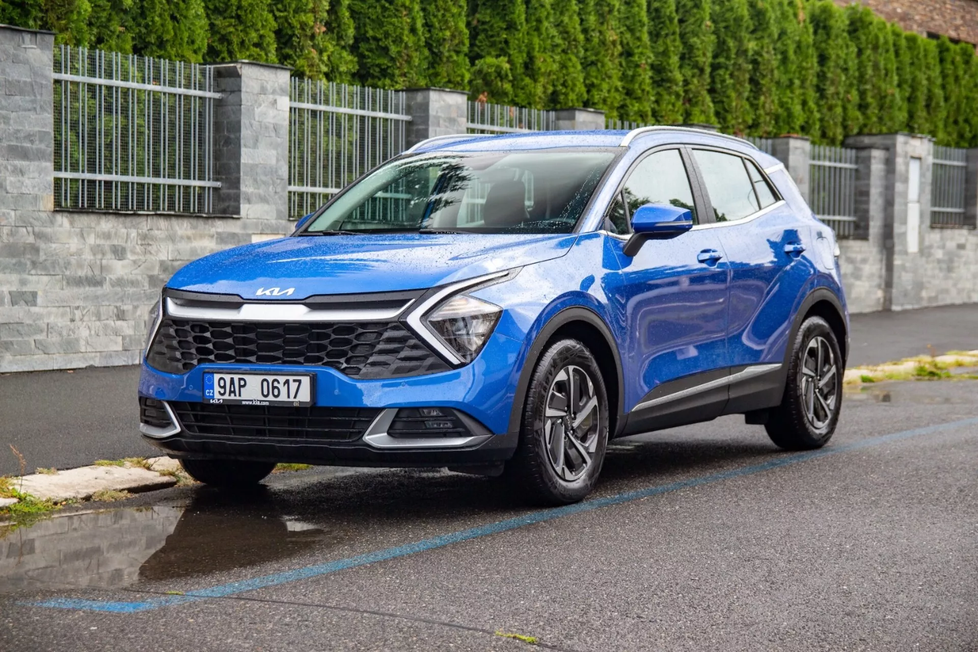 Niebieskie kompaktowe SUV Kia Sportage 2022 zaparkowane na mokrej asfaltowej ulicy pokazujące grill, reflektory, zderzak, maskę i profil strony kierowcy z trójkwartalnego przodu pod niskim kątem, aby podkreślić design pojazdu i funkcje technologiczne