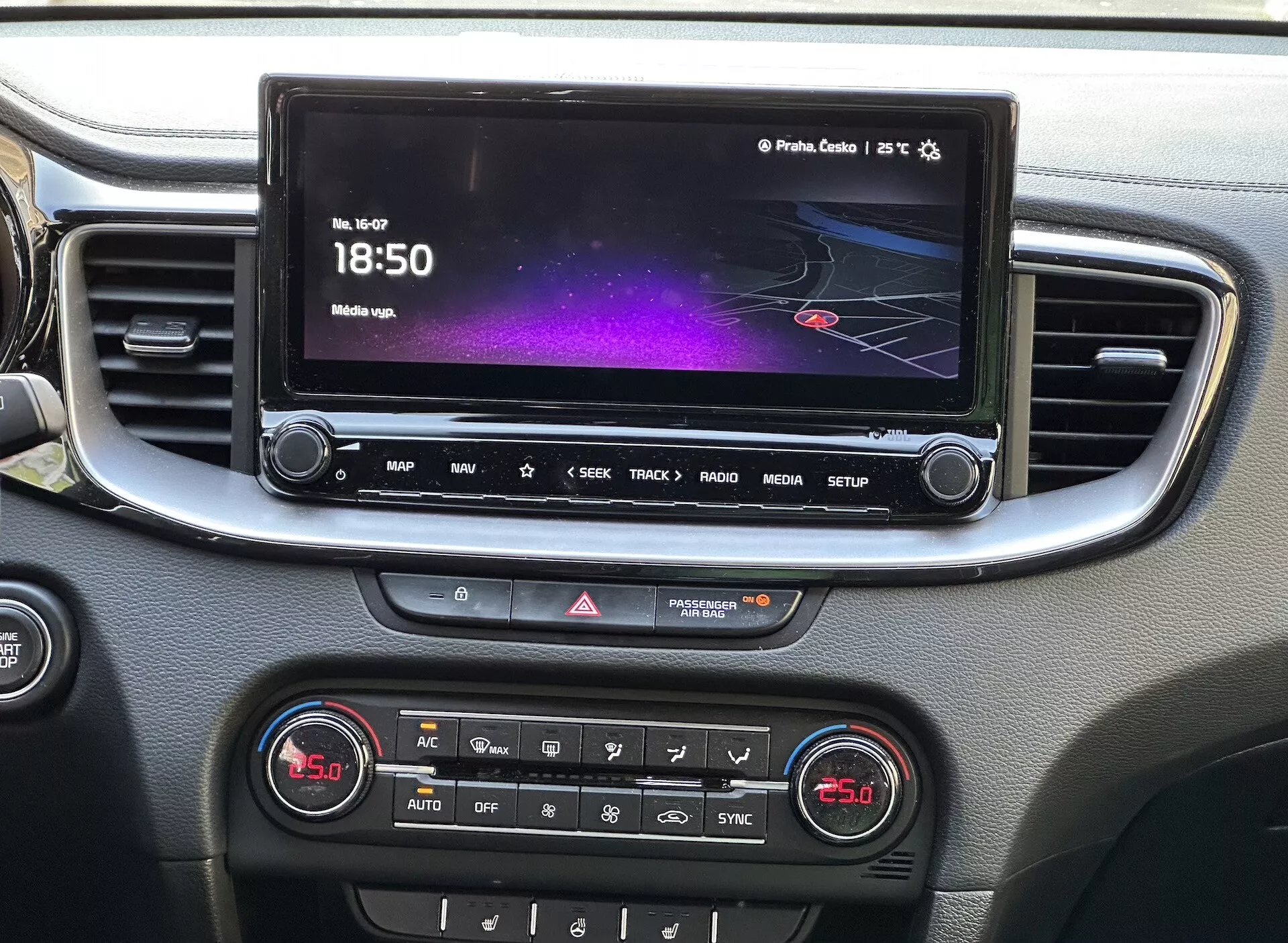 Szczegółowy widok wnętrza Kia Xceed 2022 przedstawiający konsolę środkową z dotykowym ekranem systemu infotainment pokazującym kolorowy interfejs z nawigacją i informacjami medialnymi, rząd przycisków szybkiego dostępu, kontrolę świateł awaryjnych oraz panel sterowania klimatyzacją dwustrefową, wszystko oprawione srebrnym wykończeniem.