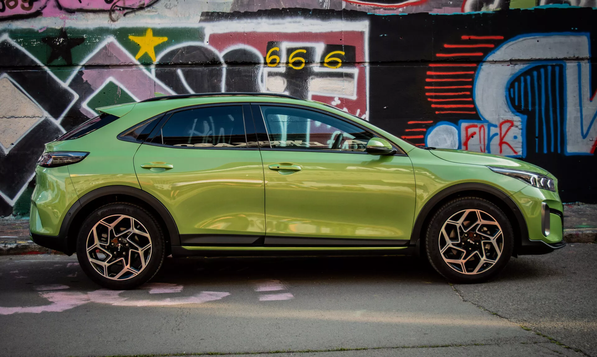 Kompaktowy SUV Kia XCeed 2022 w kolorze limonkowym zaparkowany przy ścianie pokrytej graffiti, prezentujący swój elegancki design i zaawansowane funkcje takie jak system bezkluczykowego dostępu i zintegrowane kierunkowskazy w lusterkach bocznych.