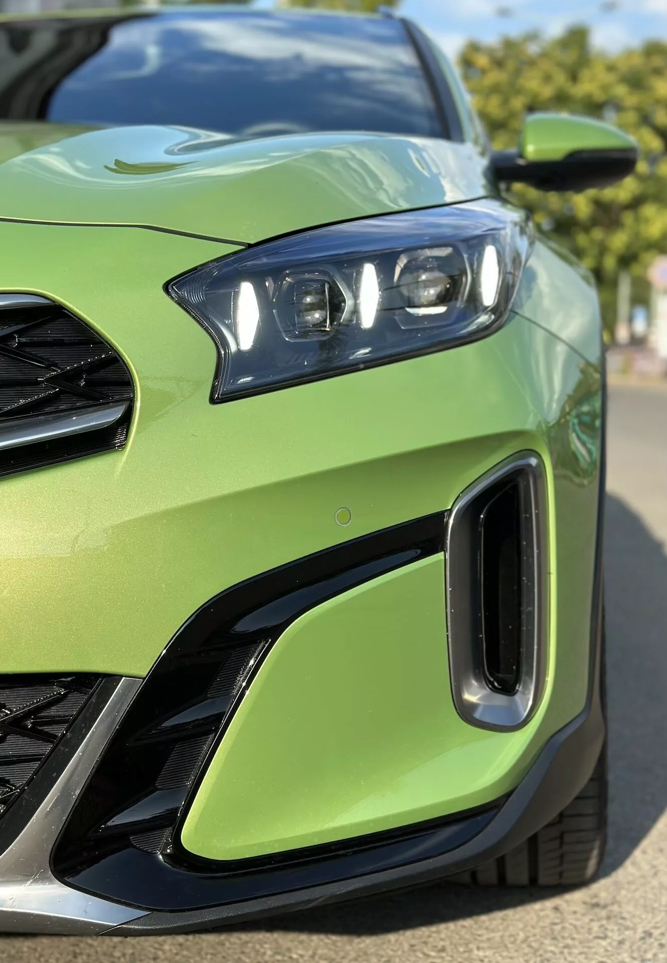Zbliżenie na przedni lewy kwadrant Kia Xceed 2022 pokazujące szczegółowo zespół reflektorów LED, część grilla, wloty powietrza na przednim zderzaku, na tle limonkowo zielonego koloru nadwozia, podkreślając sportowy design pojazdu i zaawansowaną technologię oświetlenia.