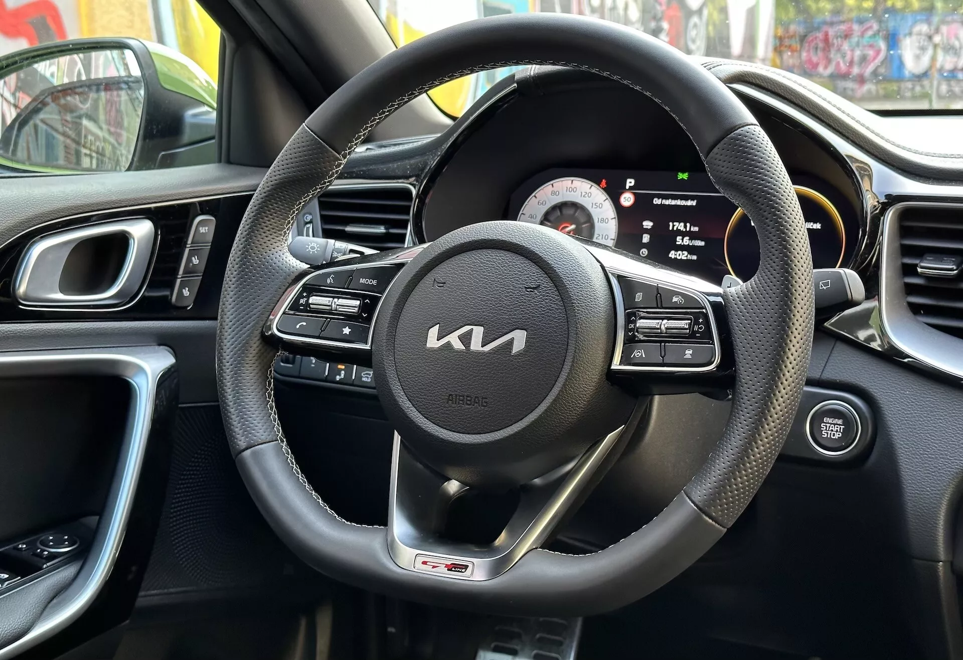 Widok wnętrza Kia XCeed 2022 z perspektywy kierowcy przedstawiający czarną kierownicę owiniętą skórą z multifunkcyjnymi kontrolami i odznaką linii GT, cyfrowo-analogowy zespół wskaźników, część konsoli środkowej z kratkami wentylacyjnymi, ergonomiczny design deski rozdzielczej, i panel drzwi kierowcy z kontrolami, odzwierciedlający miejskie otoczenie