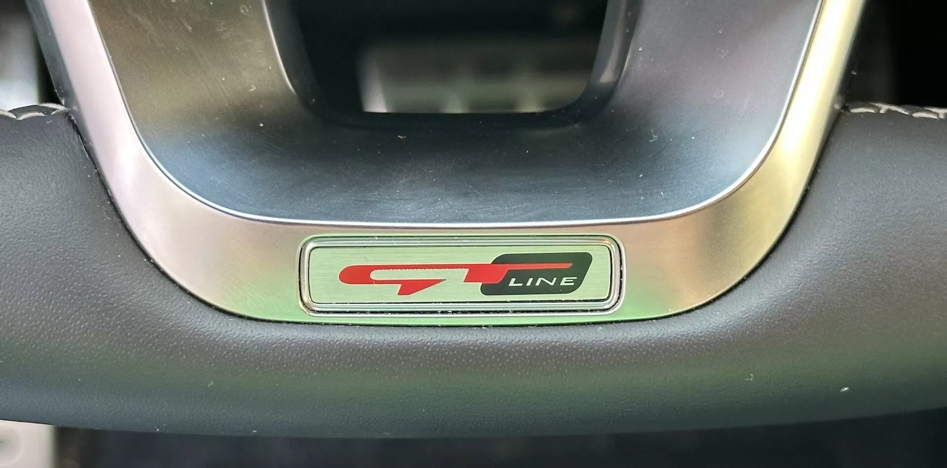 Zbliżenie na emblemat kierownicy Kia XCeed 2022 GT-Line z metalicznymi i czarnymi literami oraz sportowym czerwonym akcentem, otoczonym przez premium kierownicę skórzaną i metalową