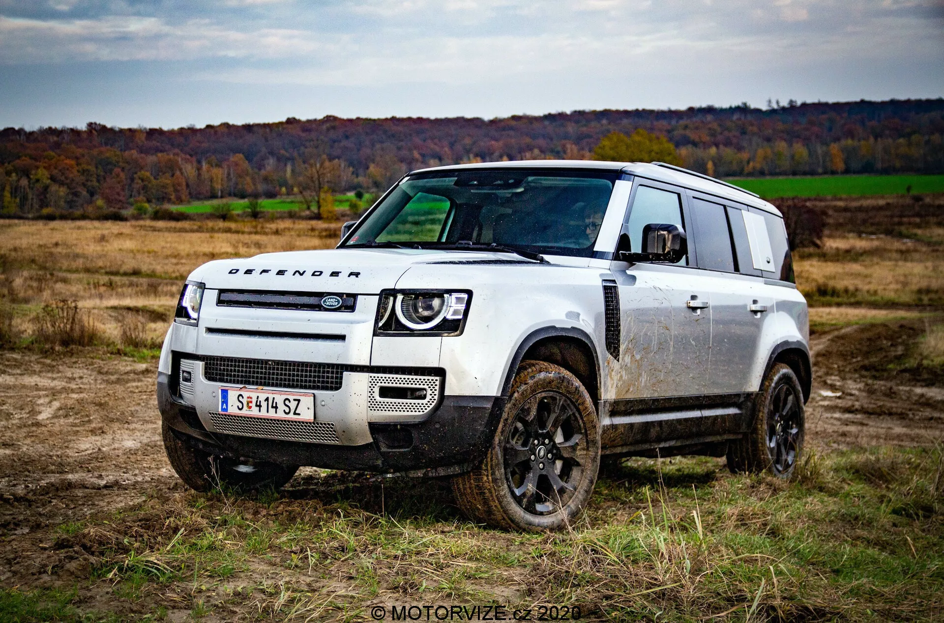 Obraz pojazdu terenowego Land Rover Defender 2020 z widokiem na trzy czwarte z przodu na otwartym polu z trawą i drzewami w tle podczas jesieni. Codzienny i kwadratowy design pojazdu, pokryty plamami błota, podkreśla jego możliwości terenowe. Znamienne cechy obejmują ciemno-kolorowe koła, napis 'DEFENDER' na masce, minimalistyczna przednia kratka z emblematem Land Rover, nowoczesne reflektory LED i solidny przedni zderzak z płytami ochronnymi. Schodkowany dach z oknami alpejskimi oraz duże tylne okna podkreślają funkcjonalny wygląd auta.