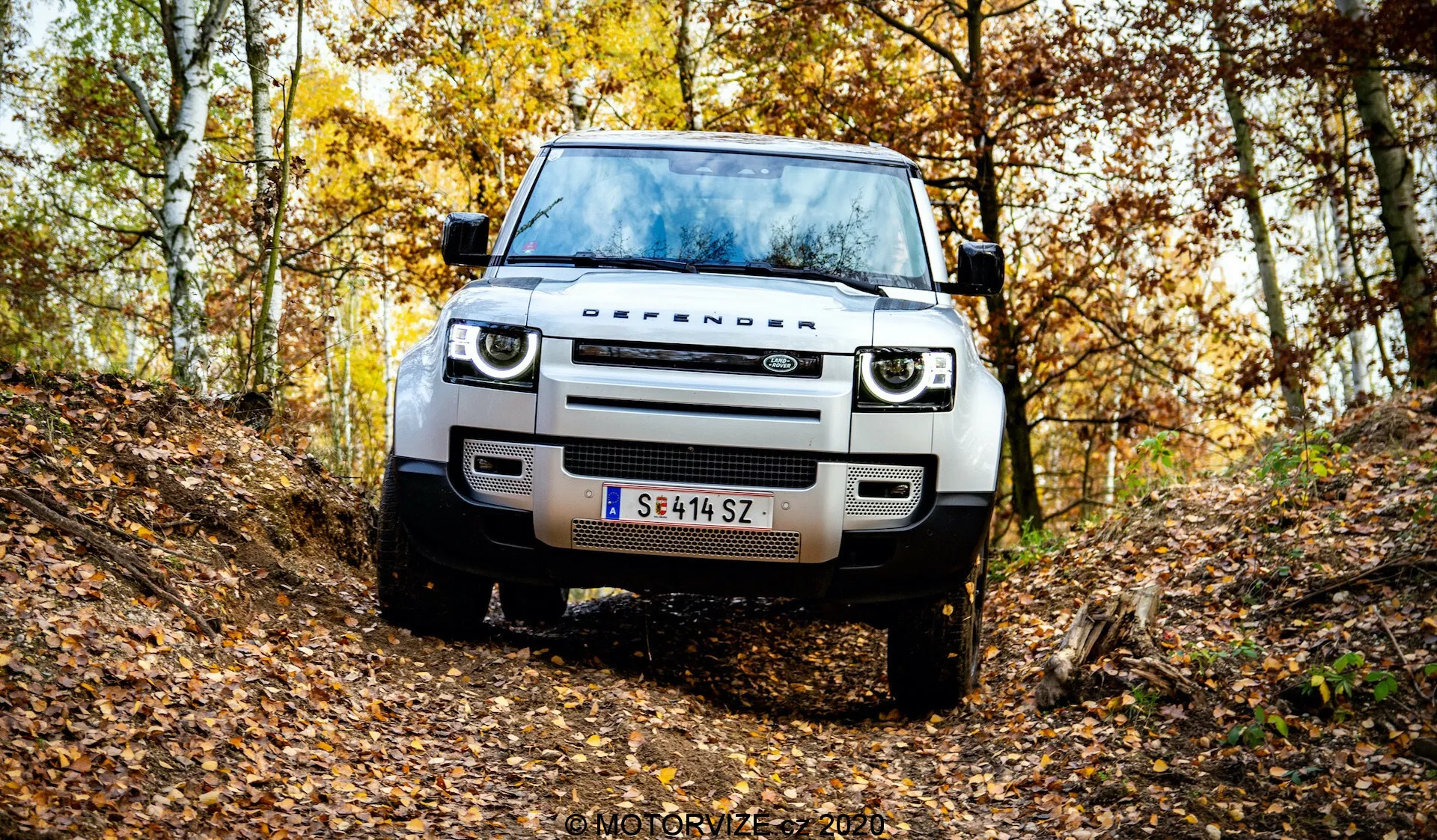 Widok z przodu nowoczesnego Land Rovera Defendera w naturalnym, jesiennym leśnym otoczeniu na nierównej ścieżce. Na zdjęciu widać grill, reflektory, maskę, koła i opony pojazdu, podkreślając jego prześwit i zdolności terenowe. Posiada nowoczesne oświetlenie LED, prosty design grilla, czujniki na przednim zderzaku lub asystent parkowania, stalową osłonę podwozia i austriacką tablicę rejestracyjną. Opadłe liście i drzewa o żółtych i brązowych liściach tworzą nastrojowy, jesienny klimat. Znak wodny 'MOTORYZE 2021' w prawym dolnym rogu.