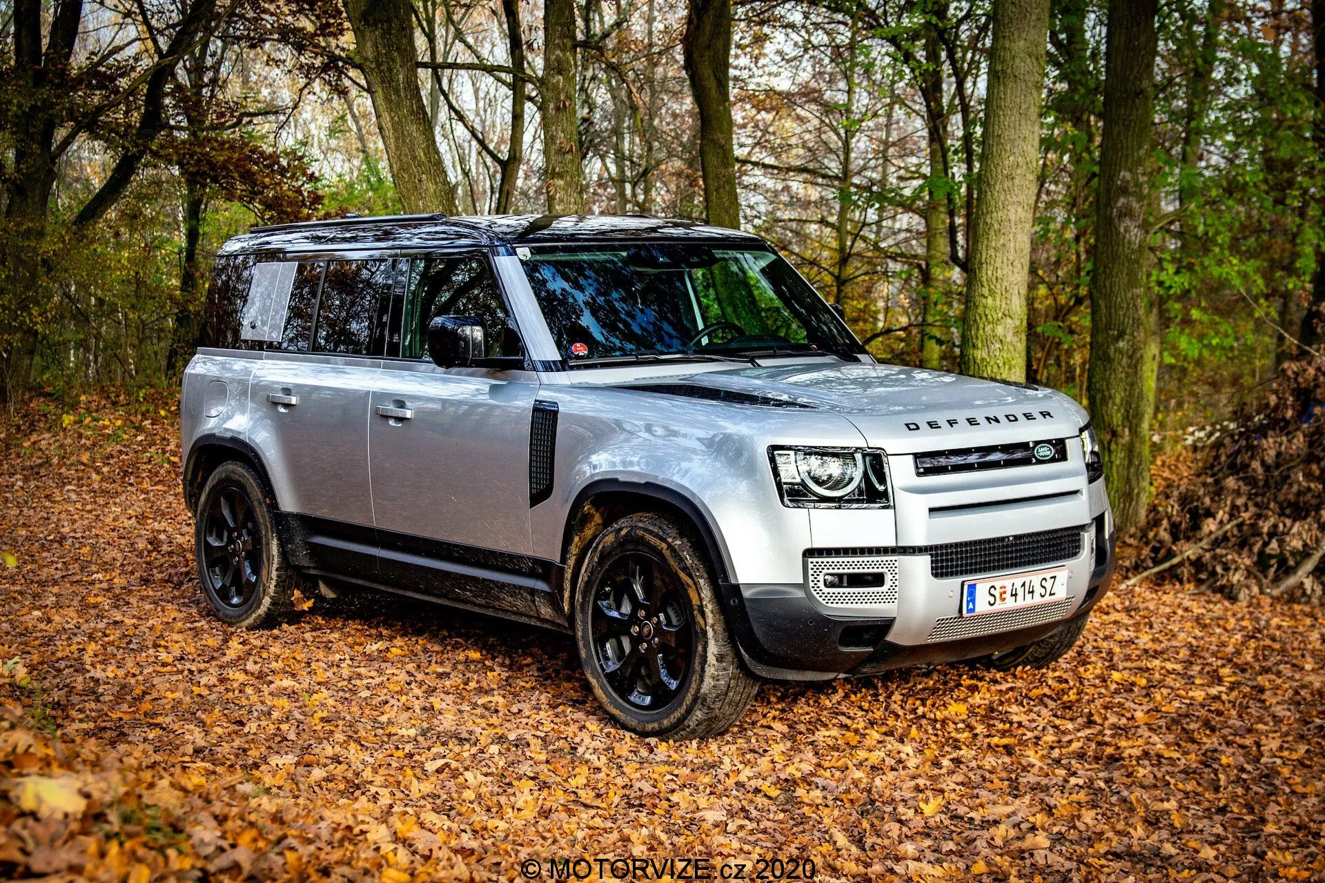 2020 Land Rover Defender w widoku trzech czwartych od przodu, ukazujący swój solidny design i zaawansowane funkcje w naturalnym, jesiennym pejzażu z opadłymi liśćmi i drzewami. SUV ma srebrne nadwozie z czarnymi detalami, reflektory LED, panoramiczny dach i czarne felgi. Obrazek uchwytuje stronę kierowcy, podkreślając elegancki grill z logo DEFENDER, czarne lusterka z wbudowanymi wskaźnikami oraz ewentualne płyty ochronne do jazdy terenowej.