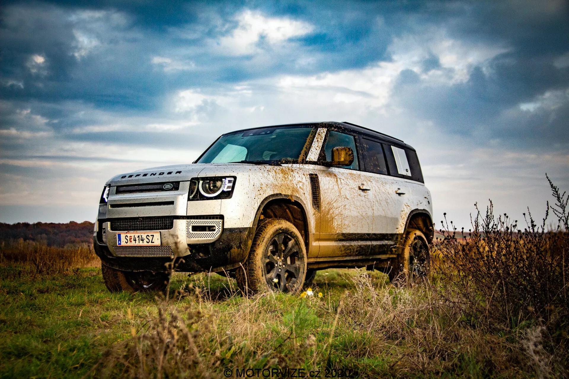 Obraz SUV Land Rover Defender 2020 na świeżym powietrzu, podkreślający jego zdolność do jazdy terenowej z perspektywy niskiego kąta. Podkreśla prześwit pojazdu, jego solidność z nadkolami, oponami i elementami ochronnymi na dolnej części nadwozia. Ukazuje widok z trzech czwartych przodu, pokazując cały przód i bok aż do tylnego drzwi, akcentując elementy designu jak reflektory, kratkę i linie konturowe na masce. Błoto i brud na panelach bocznych i kołach sugerują użytkowanie terenowe, wskazując na wytrzymałość i zdolność do jazdy terenowej. Pochmurne niebo i jesienny krajobraz w tle zwiększają poczucie przygody.