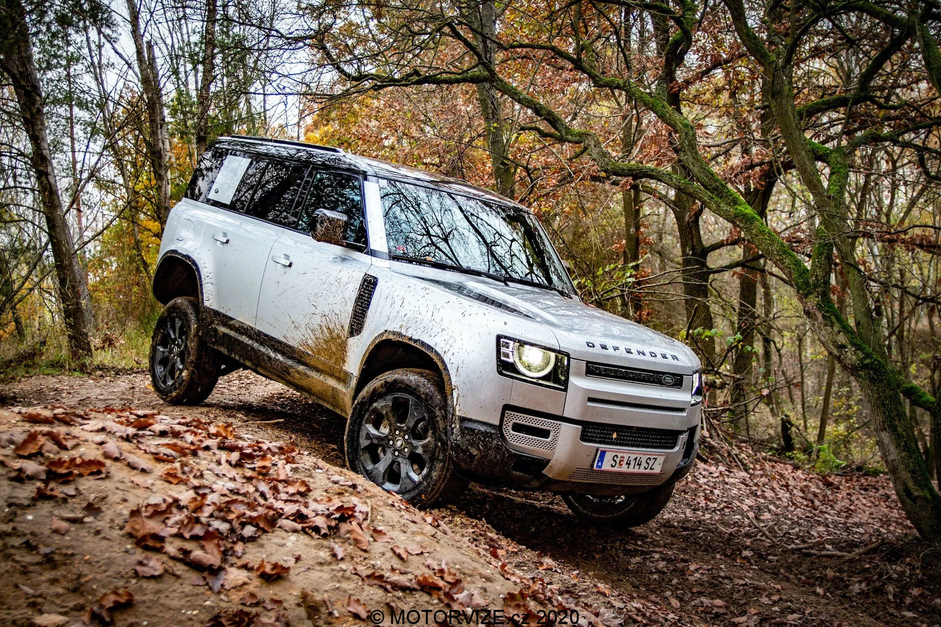 Widok z boku na białego SUV-a Land Rover Defender 2020 w off-roadowym otoczeniu; charakterystyczny design z widokiem na przedni grill i zderzak, nowoczesne reflektory LED, czarne felgi aluminiowe z terenowymi oponami, rozłożone lusterka boczne, kanciasta sylwetka z widocznym brudem, schowane klamki drzwi, płaska linia dachu i duży prześwit. Pojazd pokonuje nachylenie, ukazując swoje zdolności terenowe.