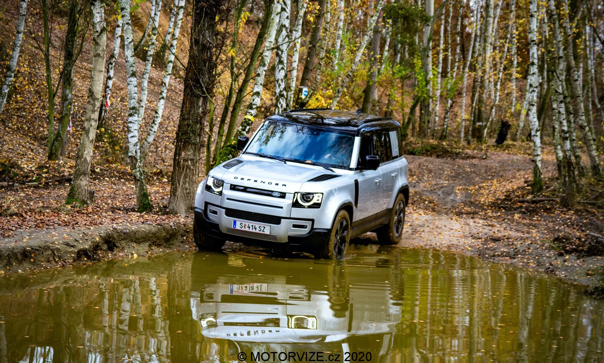 Obraz Land Rover Defender 2020 w leśnej scenerii, uchwycony pod kątem trzech czwartych z przodu. Pojazd wykazuje zdolności terenowe, jadąc przez błotnistą, wypełnioną wodą ścieżkę. Design zawiera nowoczesne reflektory, wyraźny grill i płytę ochronną. Relingi dachowe, opony z głębokim bieżnikiem i odbicie w wodzie dodają estetyki. Otoczony brzozami i opadłymi liśćmi, obraz uchwytuje istotę jesieni.