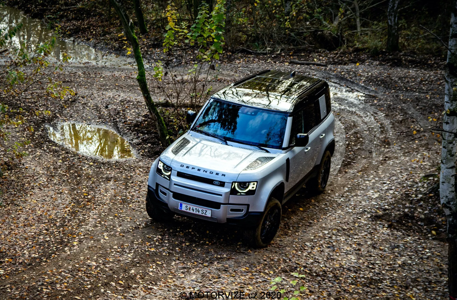 Widok z lotu ptaka na Land Rover Defender 2020 poruszającego się po off-roadowym szlaku, z widocznymi szczegółami takimi jak przedni grill, maska, przednia szyba i część strony kierowcy, otoczony naturalnym otoczeniem z kałużami wody i błota oraz drzewami.