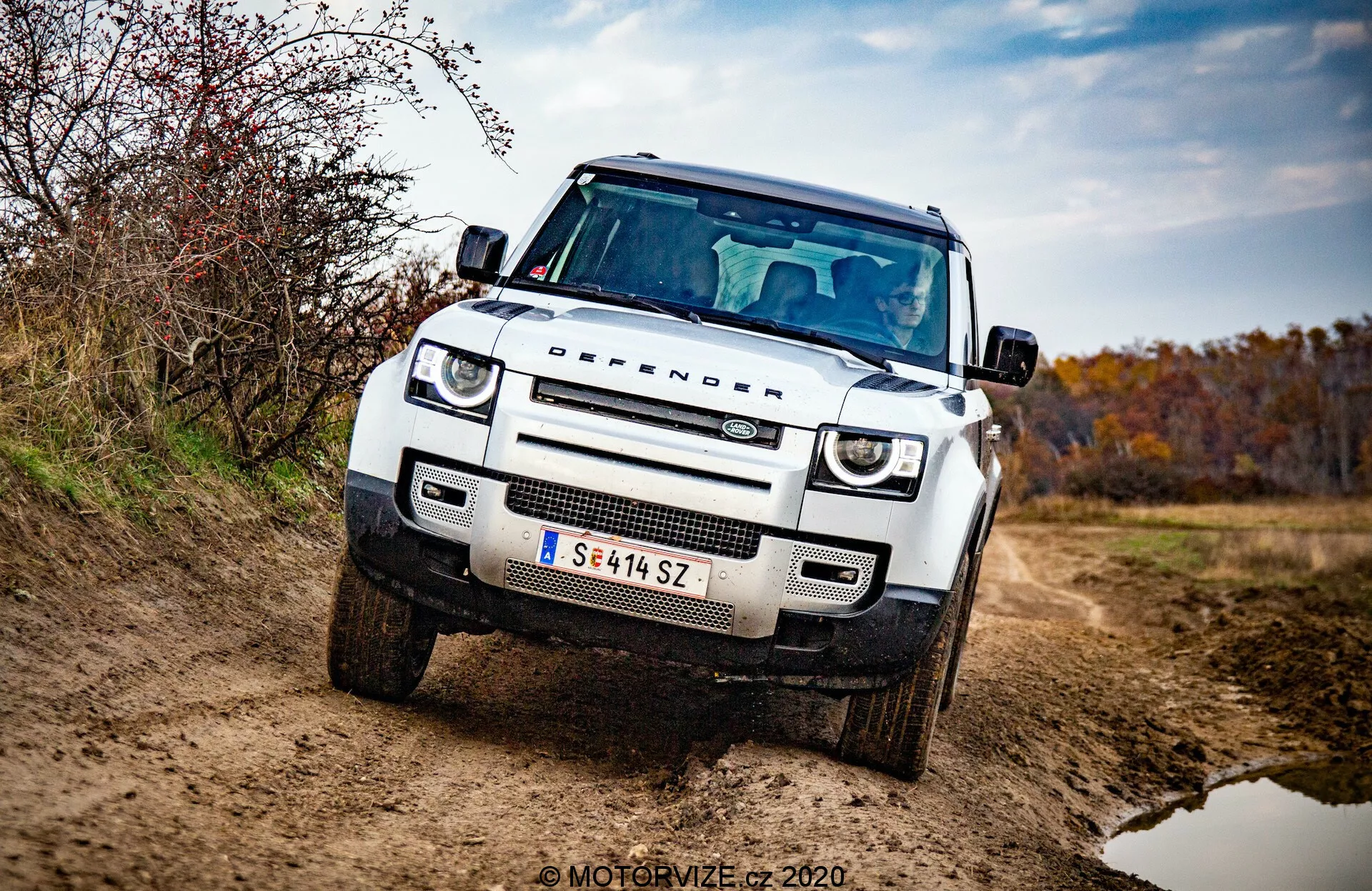 Land Rover Defender 2020 z przodu w widoku trzech czwartych, biały, pudełkowaty SUV terenowy z płaską maską, kwadratowymi nadkolami, okrągłymi światłami do jazdy dziennej LED, reflektorami adaptacyjnymi, kratką z siatki plastra miodu z logo marki, płytą ochronną na przednim zderzaku, podwyższoną i solidną postawą, lusterkiem bocznym z wbudowanymi kierunkowskazami i naciskiem na kratkę, reflektory i przedni zderzak. Pojazd na nieutwardzonej drodze z brudem na zewnętrznej stronie, otoczony ziemistym, jesiennym środowiskiem. Profesjonalne zdjęcie dobrze oświetlone z rozmytym tłem i znakiem wodnym 'MOTORVIZE © 2020'.