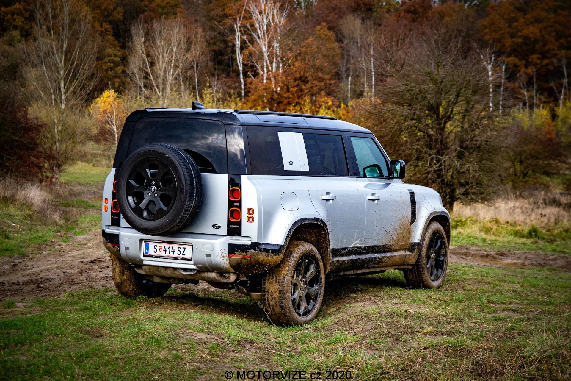 Trzyczwarte tylne ujęcie nowoczesnego Land Rover Defender 2020, czterodrzwiowego SUV-a zaparkowanego na trawiastej okolicy z widocznymi śladami błota na bokach, wskazującymi na niedawne używanie poza drogami. Zdjęcie z poziomu ziemi podkreśla profile tylne i boczne, koncentrując się na tylnym projekcie, w tym widocznych światłach tylnych, tylnym kole, obszarze ładunkowym i kole zapasowym zamontowanym na tylnej klapie z logo marki. Pojazd prezentuje dwukolorowy schemat kolorów z jasnym nadwoziem i czarnym dachem, aluminiowe felgi z masywnymi oponami terenowymi, dużym prześwitem i częściowo zasłoniętym kołem zapasowym tylnym oknem oraz tylną wycieraczką. Błotniste boki i dolne panele sugerują, że SUV był prowadzony po błotnistym terenie. W tle można dostrzec siedlisko leśne lub zalesione z miękkim światłem, wskazującym na pochmurne niebo lub warunki słabego oświetlenia.