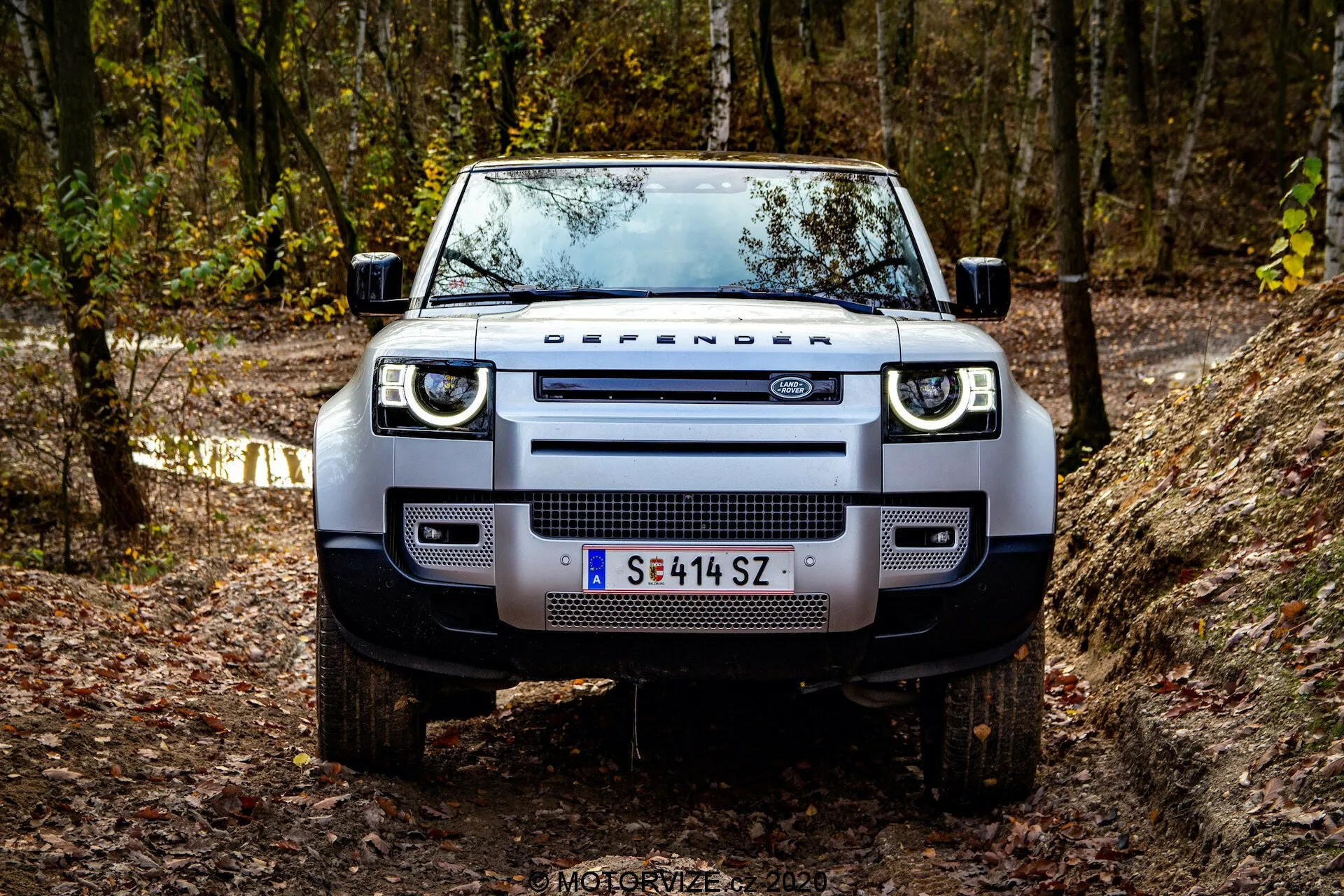 Widok z przodu Land Rovera Defender 2020 na szlaku off-road, ukazujący jego solidną i dominującą obecność. Widoczne szczegóły obejmują charakterystyczny grill z napisem 'DEFENDER', nowoczesne prostokątne reflektory z LED-owymi światłami do jazdy dziennej, szeroki przedni zderzak z zintegrowanymi światłami przeciwmgielnymi, solidne osłony podwozia, płaską maskę z wyraźnymi krawędziami, masywne opony terenowe na felgach aluminiowych oraz funkcjonalne lusterka boczne z zintegrowanymi kierunkowskazami. Pojazd znajduje się w zalesionym terenie z jesiennym listowiem, co podkreśla jego terenowy i przygodowy charakter.
