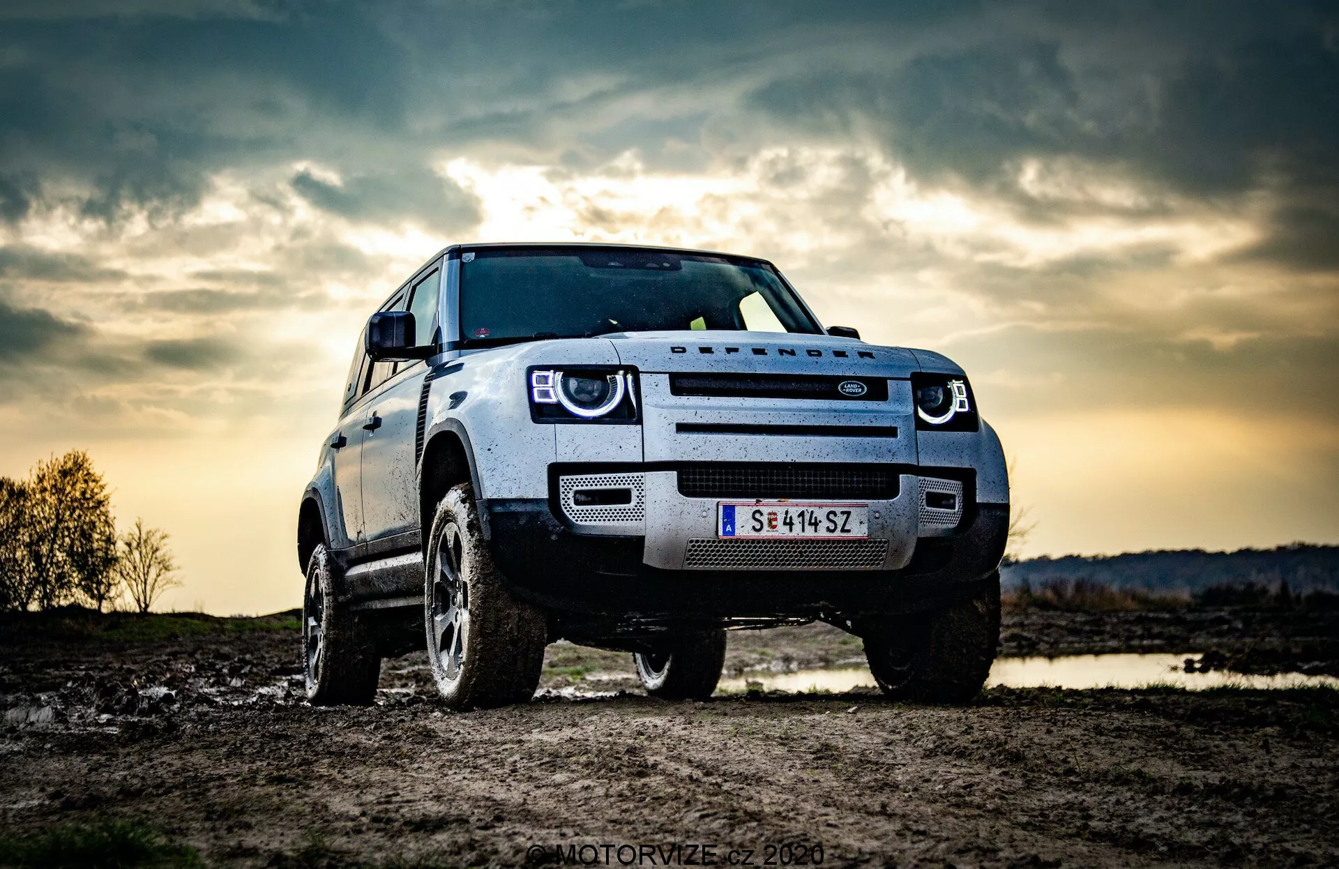 Obraz przedstawia SUV Land Rover Defender 2020 zaparkowany na zewnątrz, prawdopodobnie w terenie. Uchwycony z perspektywy niskiego kąta, podkreśla solidną sylwetkę i wysokość pojazdu. Widok przednich trzech czwartych pokazuje przedni grill, reflektory, maskę i błotnik oraz koło po stronie kierowcy. Widoczne są charakterystyczne diodowe światła do jazdy dziennej, co sugeruje nowoczesne funkcje oświetleniowe. Wyrazisty przedni grill prawdopodobnie odzwierciedla tożsamość producenta, a przednia kamera lub czujnik na grillu wskazuje na zaawansowane systemy wspomagania kierowcy. Dramatyczne tło nieba, prawdopodobnie przy wieczornym lub zmierzchowym oświetleniu, dodaje poczucia przygody, podczas gdy solidne koła i duży prześwit podkreślają zdolność do jazdy w terenie.
