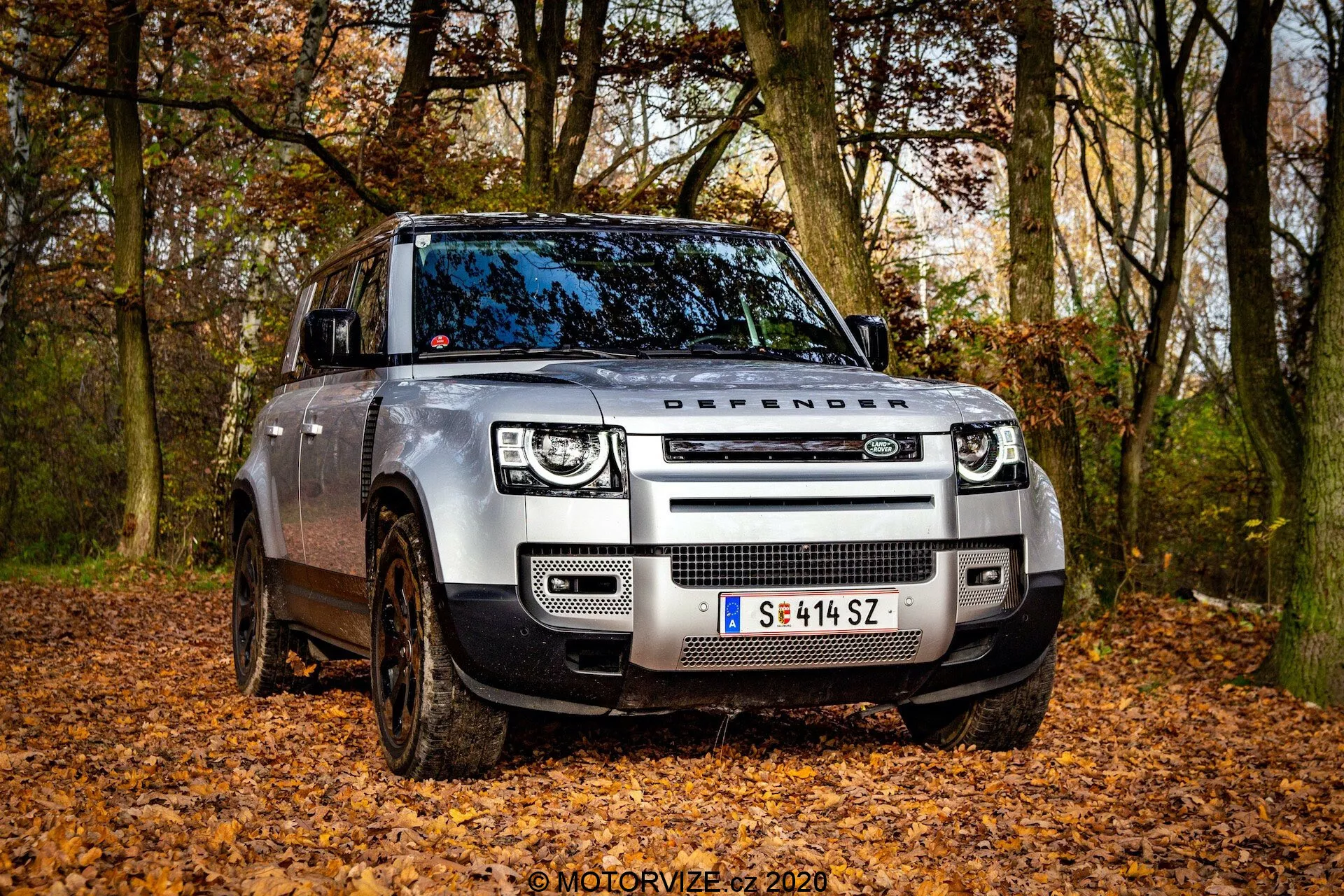 Widok z przedniego kąta nowoczesnego Land Rover Defender 2020 w terenowym otoczeniu z opadłymi liśćmi, uchwycony z niskiej perspektywy. Obraz podkreśla przednią kratkę z napisem 'DEFENDER' na masce, reflektory LED z kwadratowymi światłami dziennymi oraz nowoczesne czujniki zintegrowane z przednim zderzakiem. Błotniste koła i regulowane zawieszenie pneumatyczne wskazują na terenowe możliwości pojazdu. Widoczna jest strona kierowcy, przednie drzwi i część tylnych drzwi, ukazując solidny design pojazdu z częściowo skręconymi kołami.